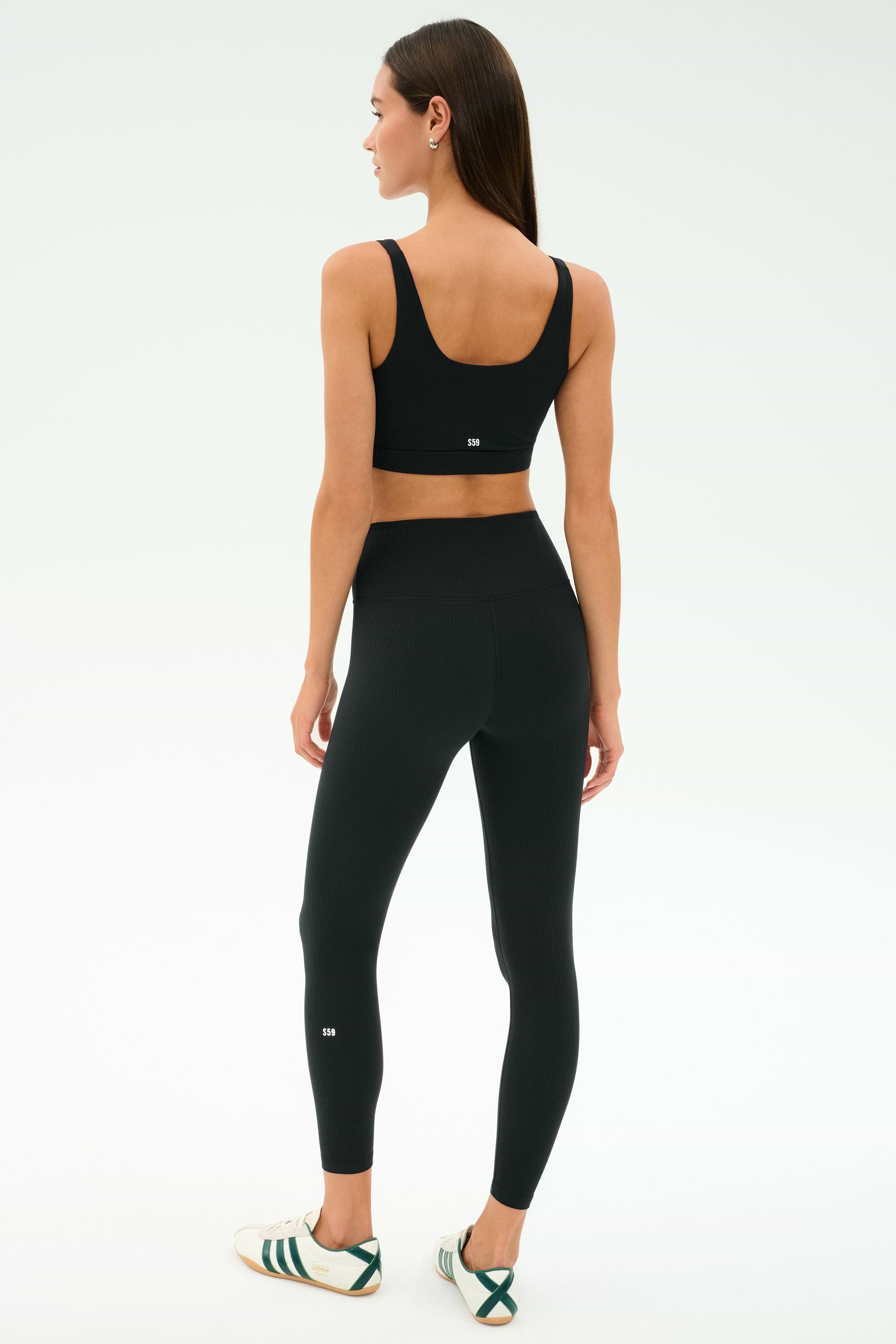Woman faces away in SPLITS59 Airweight Rib High Waist 7/8 leggings (Black) and a black sports bra, paired with white sneakers featuring green stripes—perfect for hot yoga or Pilates—set against a plain light background.
