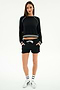 A woman stands facing forward wearing the SPLITS59 Brooks Fleece Sweatshirt With Stripe in black/white, paired with black shorts featuring a white drawstring, white socks, and white sneakers against a plain white background.