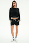 A woman stands facing forward wearing the SPLITS59 Brooks Fleece Sweatshirt With Stripe in black/white, paired with black shorts featuring a white drawstring, white socks, and white sneakers against a plain white background.