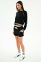 A woman wears SPLITS59's Reena Fleece Sweatshort in black—stretchy material with a white drawstring—paired with a matching black long-sleeve top with white trim, white socks, and sneakers, all set against a plain white background.