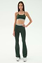A woman faces forward, wearing the SPLITS59 Raquel Active Rib Flare in Heather Military—dark green high-waisted leggings and matching sports bra with white stripes—paired with white sneakers, against a plain light background.