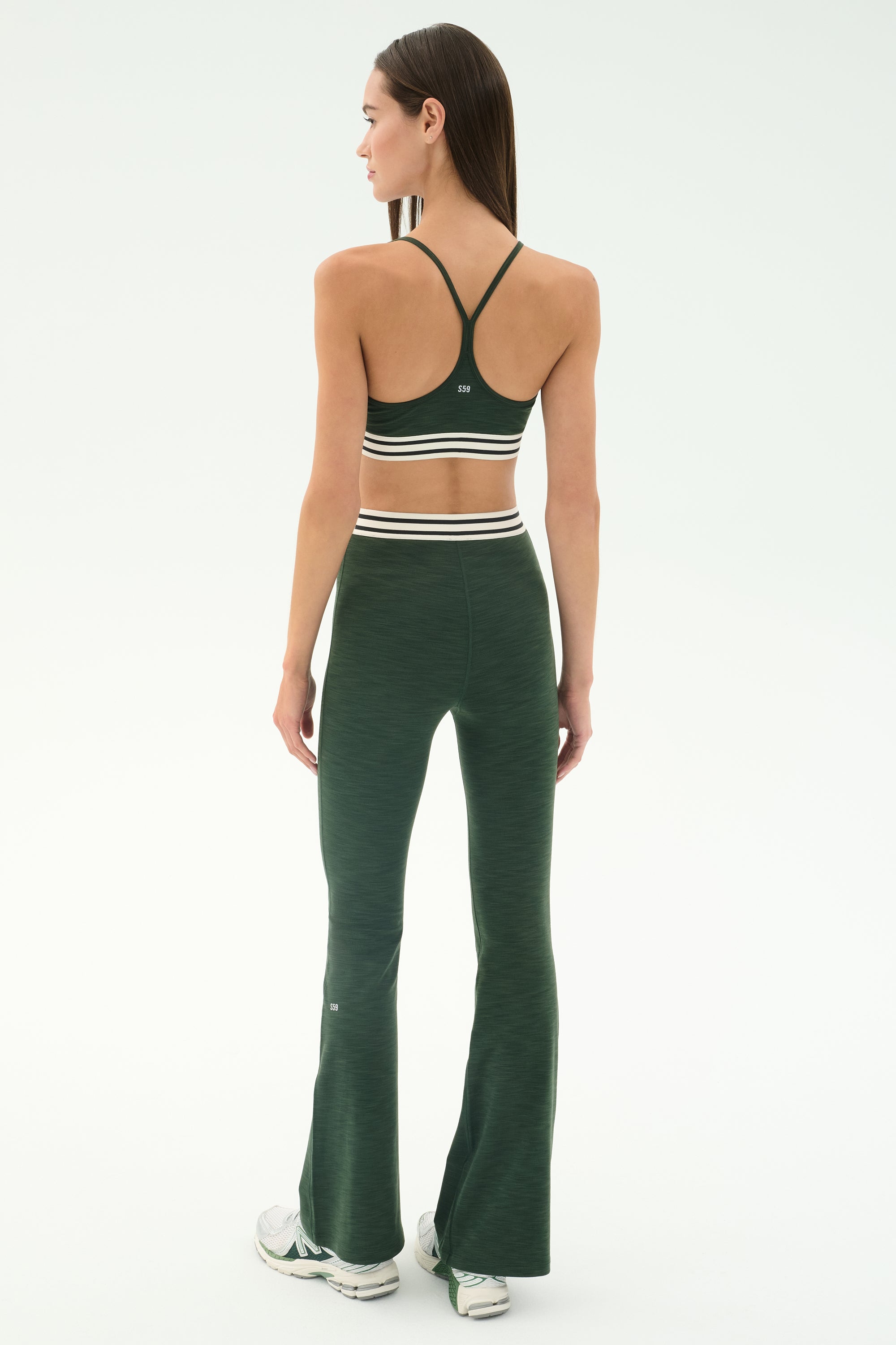 A woman faces away in the SPLITS59 Raquel Active Rib Flare - Heather Military set, featuring a dark green spandex sports bra and matching high-waisted flared leggings, paired with white sneakers against a plain light background.