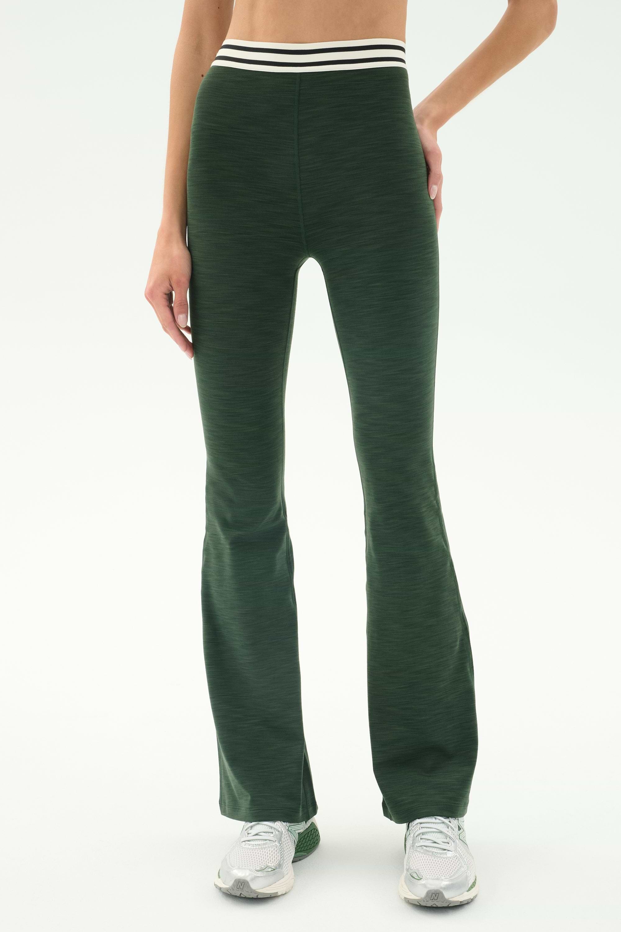 A person wears SPLITS59's Raquel Active Rib Flare - Heather Military pants with a white and black striped waistband and white sneakers, standing against a plain light background.