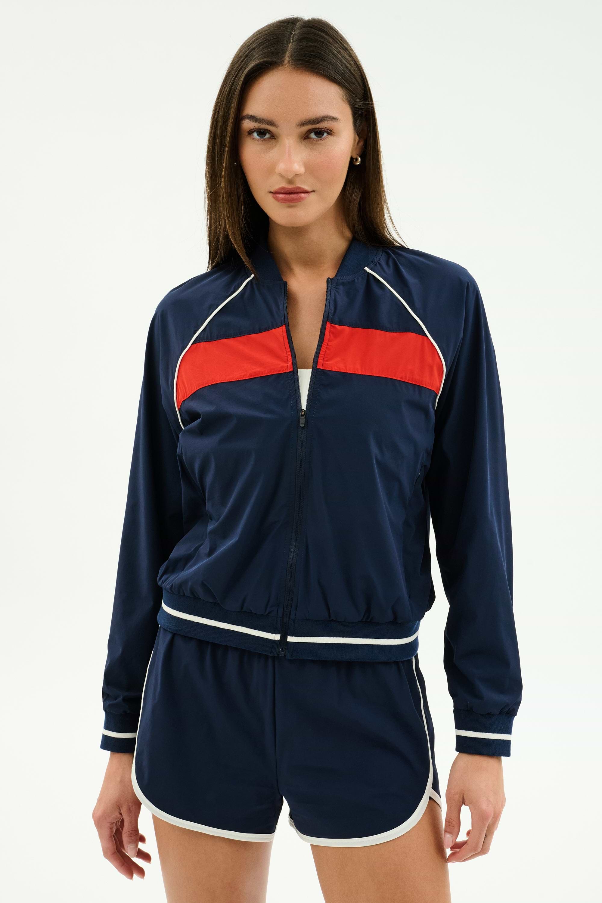 A woman faces forward wearing the SPLITS59 Carrie Woven Nylon Jacket in Indigo/Lotus Red, paired with matching navy blue spandex shorts featuring white trim.
