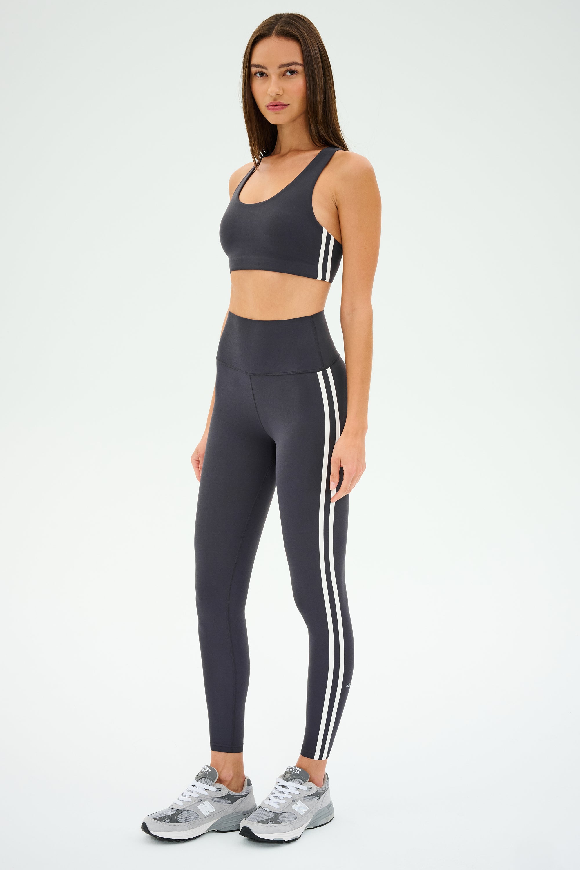 In athletic wear against a plain backdrop, the individual showcases a SPLITS59 Ella Airweight Bra in Graphite/White and leggings with white stripes, complemented by gray sneakers—perfect for hot yoga or simply enjoying the ultra-luxe Airweight experience.