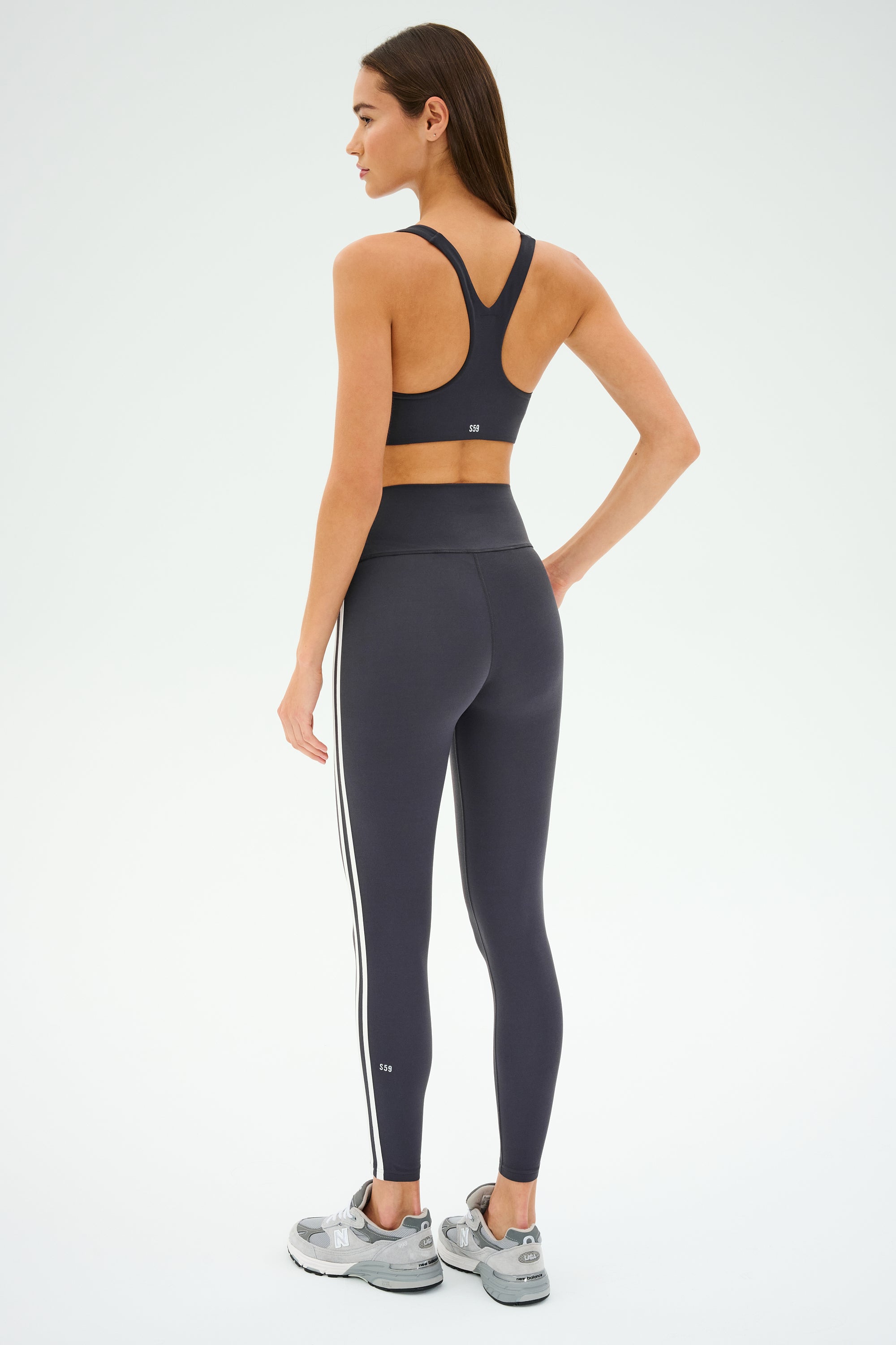 A person showcases the SPLITS59 Ella Airweight Bra in Graphite/White, paired with matching leggings and gray sneakers, on a white background. Perfect for hot yoga, this chic set blends style and function seamlessly.