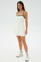 A person stands wearing the SPLITS59 Monah Rigor Dress in White/Arugula, featuring green straps, paired with white sneakers against a plain white background.