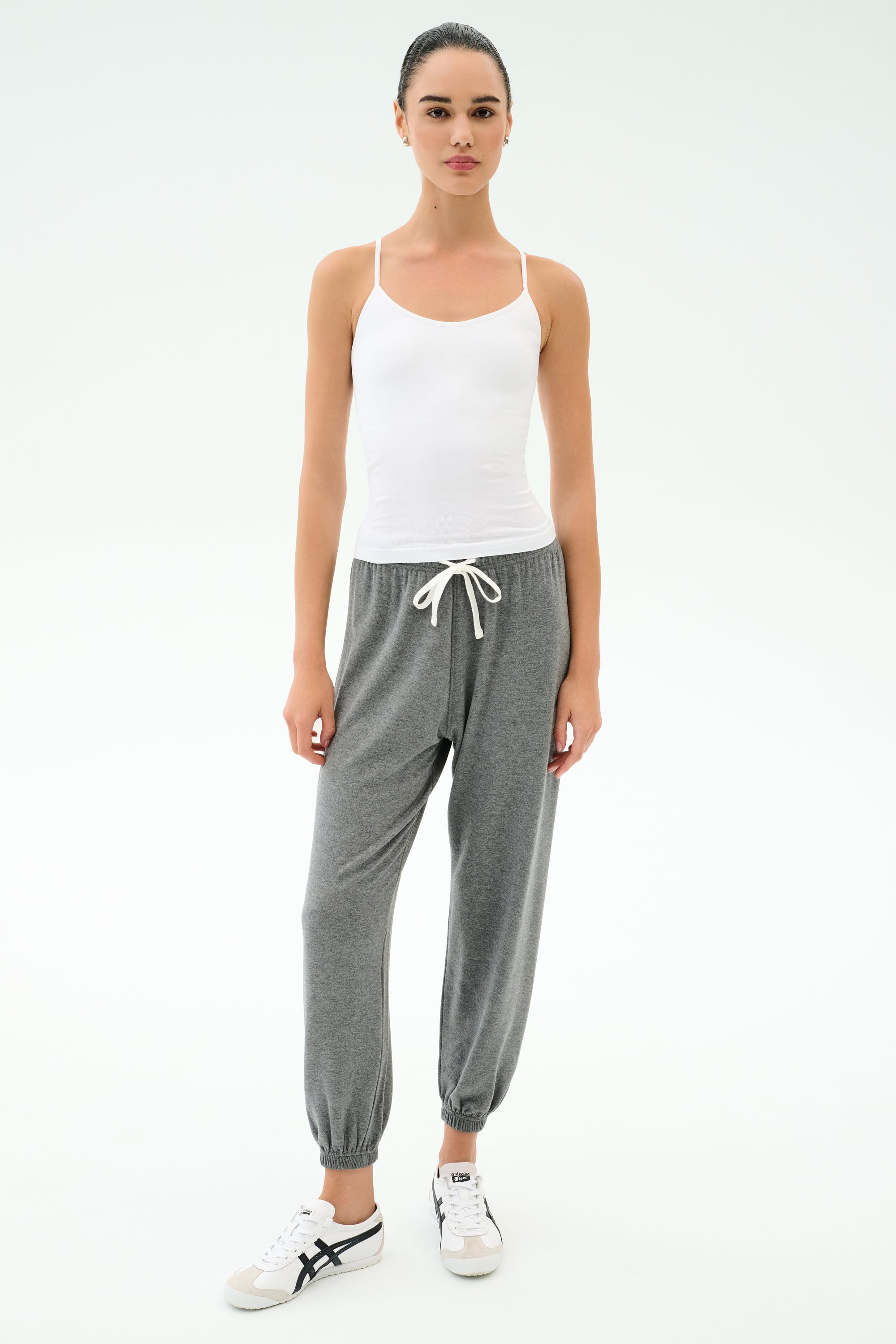 A woman faces forward on a white background, wearing the SPLITS59 Loren Seamless Waist Length Tank in white, paired with gray drawstring jogger pants and white sneakers.
