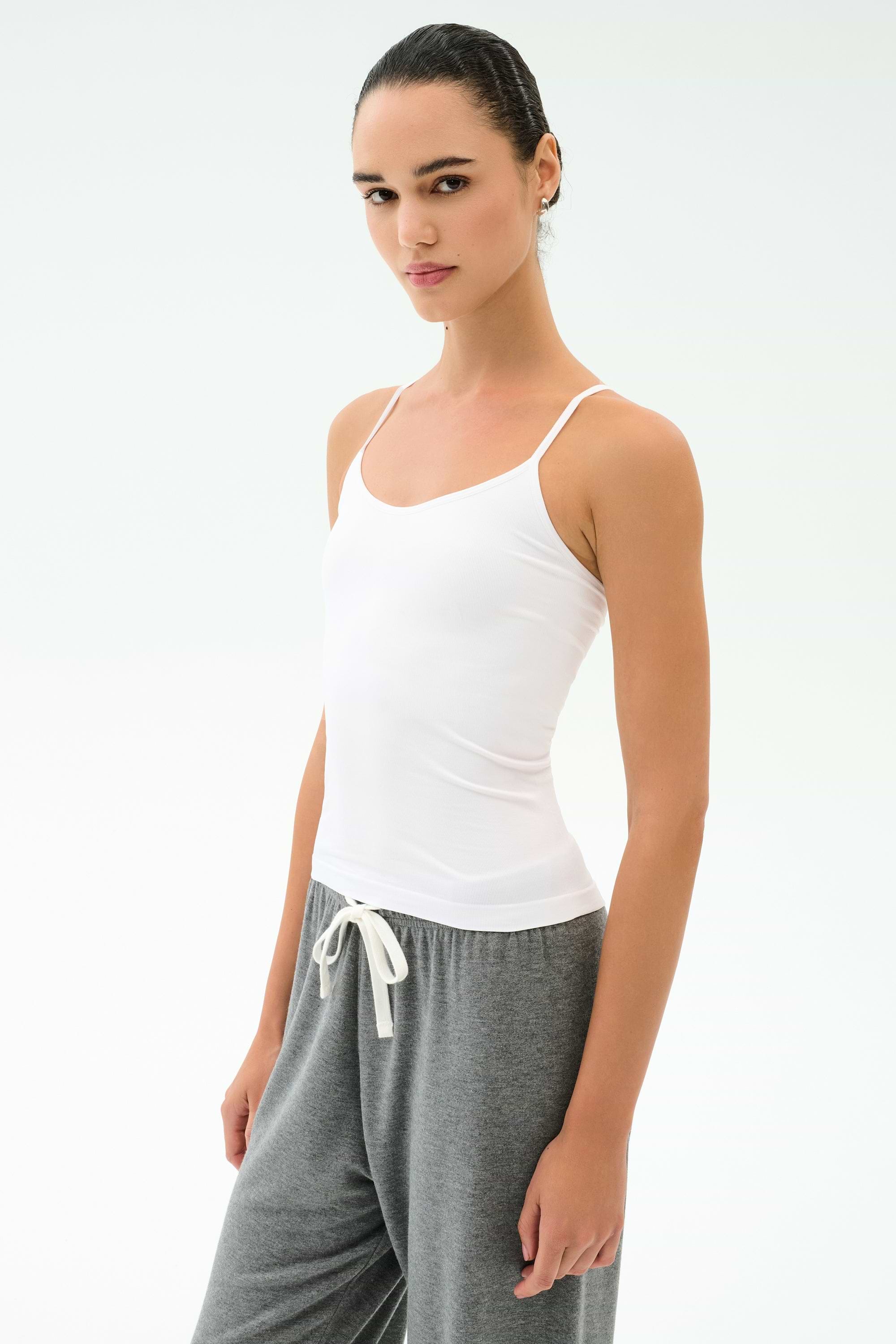 A person stands slightly sideways against a plain background wearing the SPLITS59 Loren Seamless Waist Length Tank in white and gray drawstring pants.