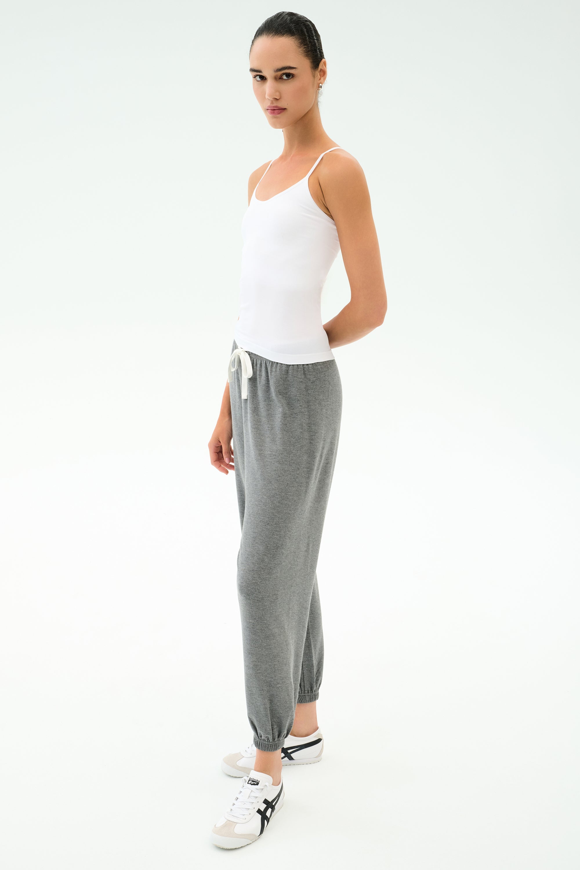 Person standing against a plain background, wearing the SPLITS59 Loren Seamless Waist Length Tank in white, gray drawstring sweatpants, and white sneakers with black accents.