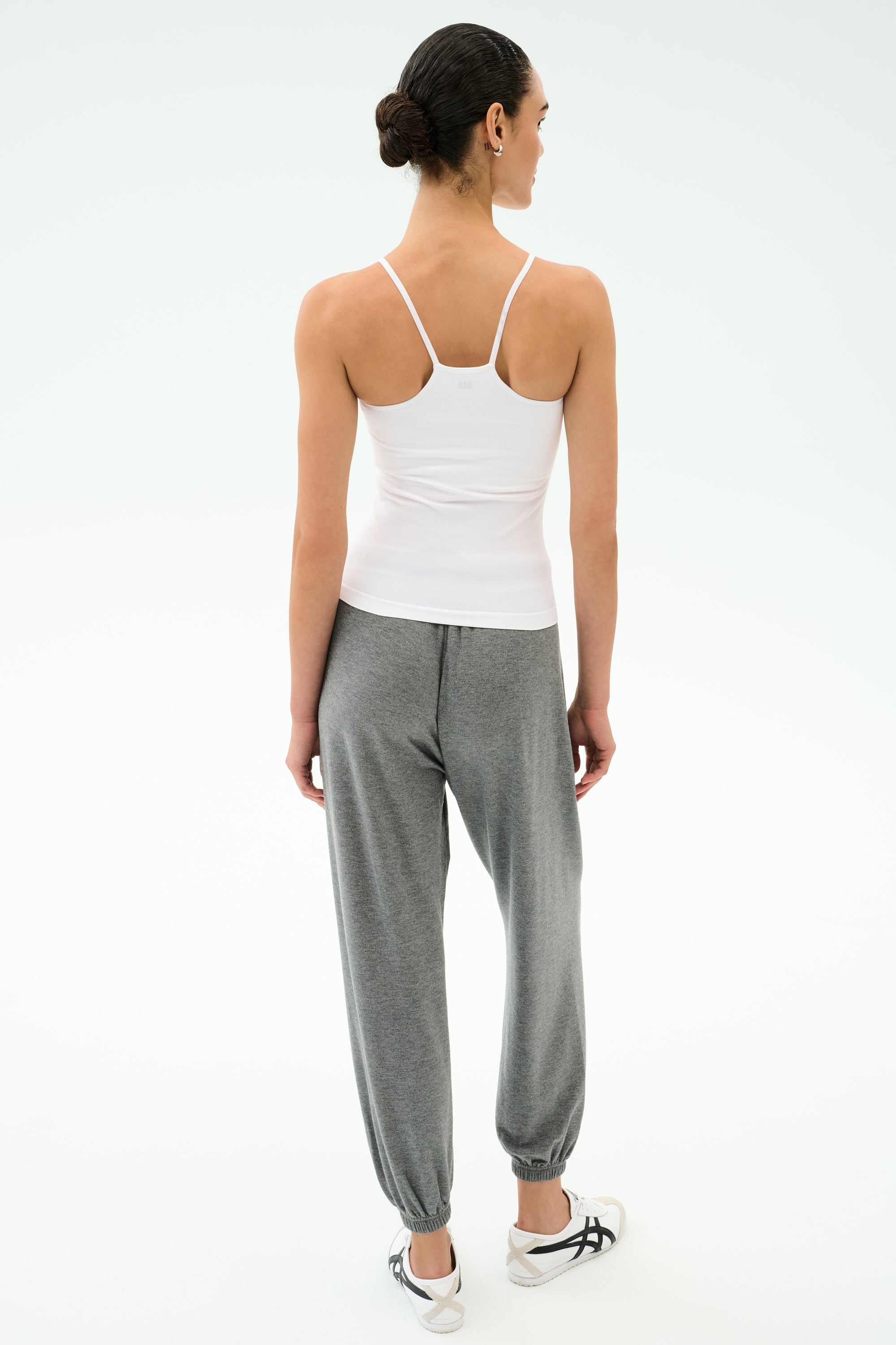 A woman faces away, wearing the SPLITS59 Loren Seamless Waist Length Tank in white, paired with gray joggers and white sneakers, set against a plain light background.