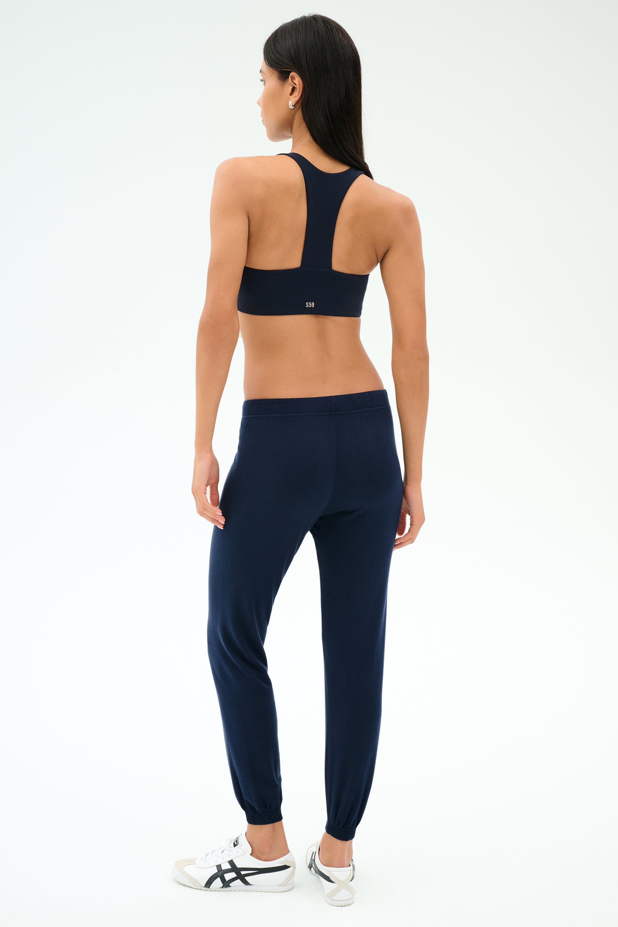 A woman faces away, wearing SPLITS59's Sonja Fleece Sweatpant in Indigo, a navy sports bra, and white sneakers against a plain light background.