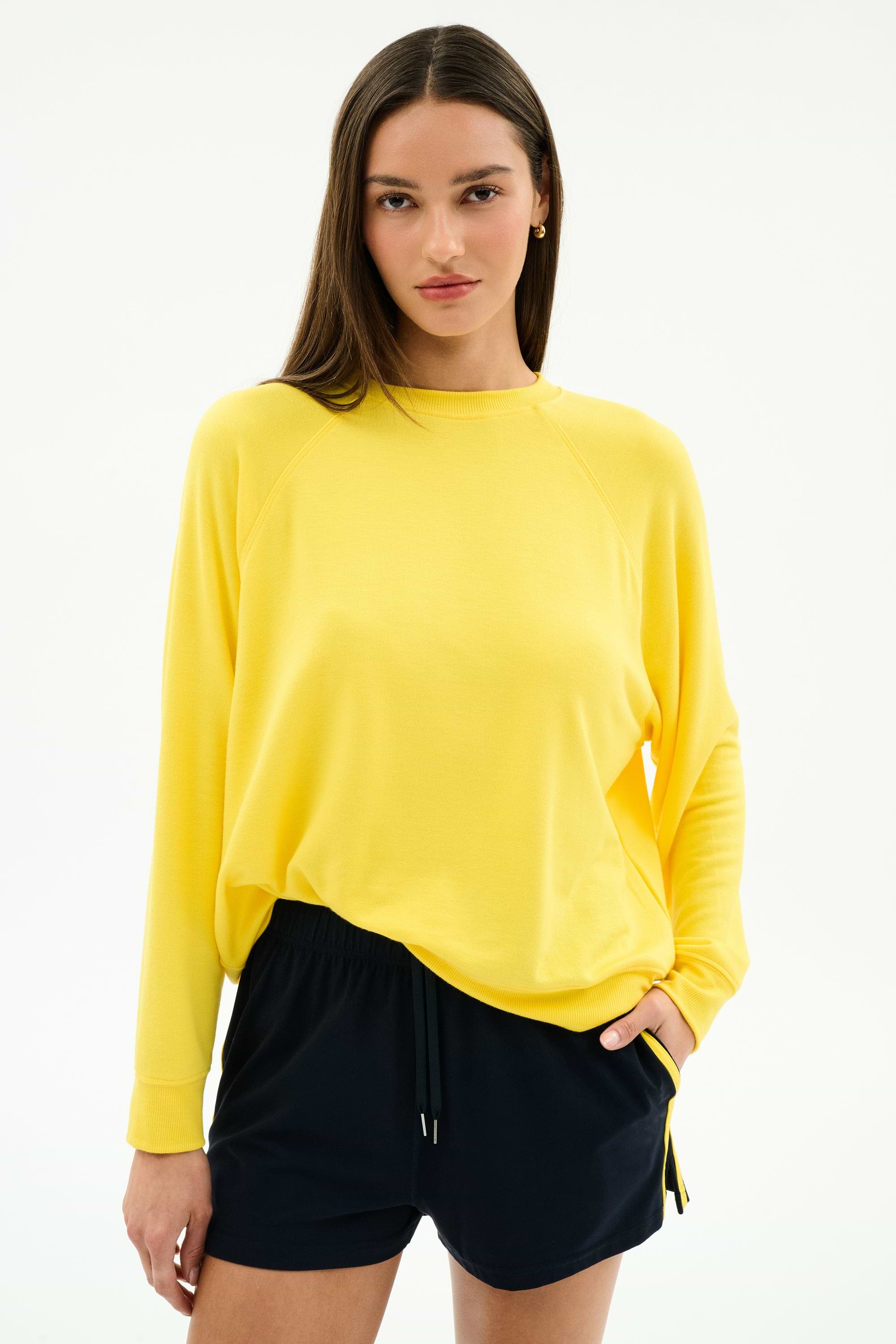 A woman stands against a plain background wearing the SPLITS59 Andie Oversized Fleece Sweatshirt in Bright Yellow, paired with black drawstring shorts, with one hand in her pocket.