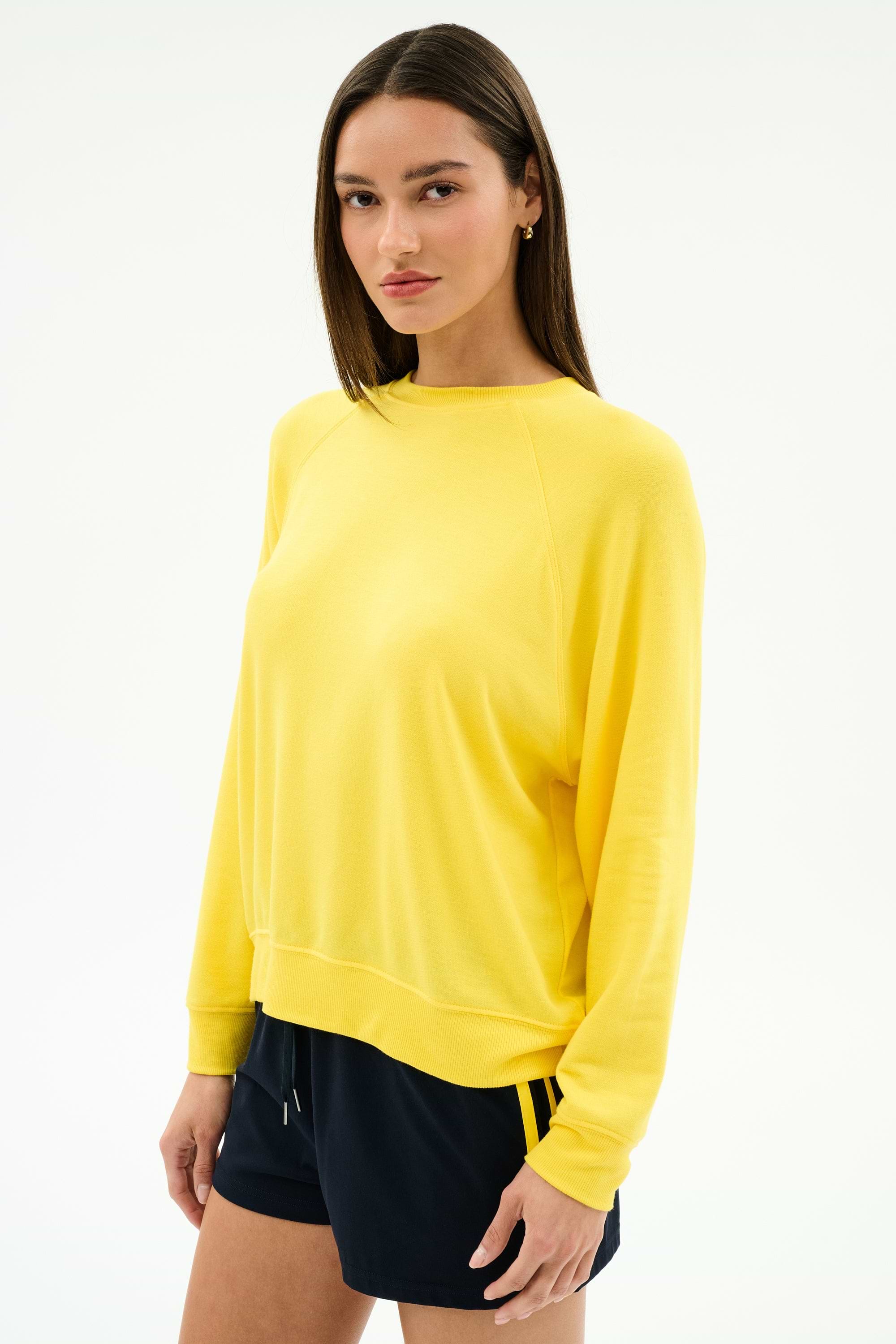 A woman stands against a white background wearing the SPLITS59 Andie Oversized Fleece Sweatshirt in bright yellow, paired with black spandex-blend shorts.