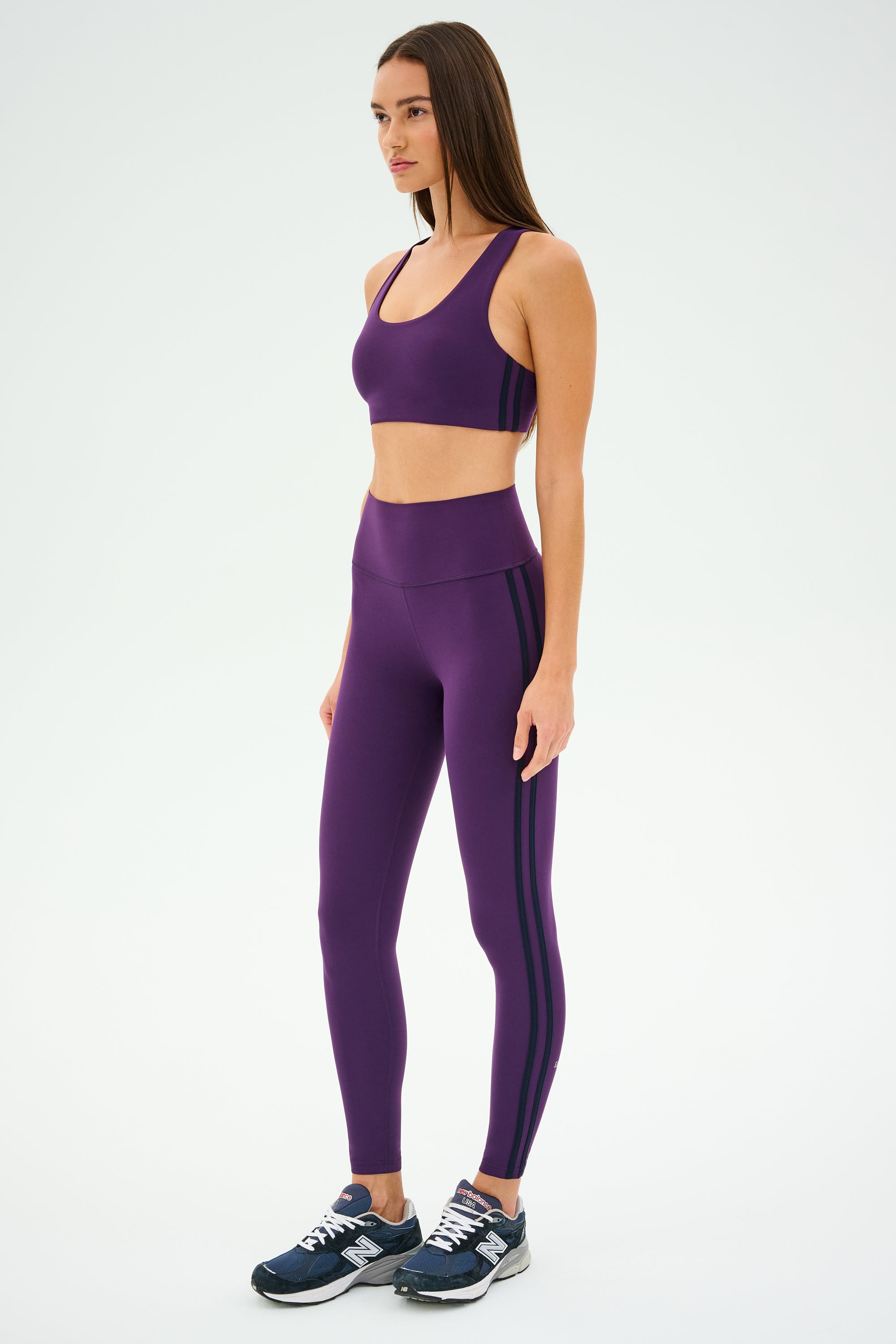 A woman in purple athletic wear, featuring a SPLITS59 Ella Airweight Bra in Black Currant/Indigo, and blue sneakers stands on a plain background, looking to the side.