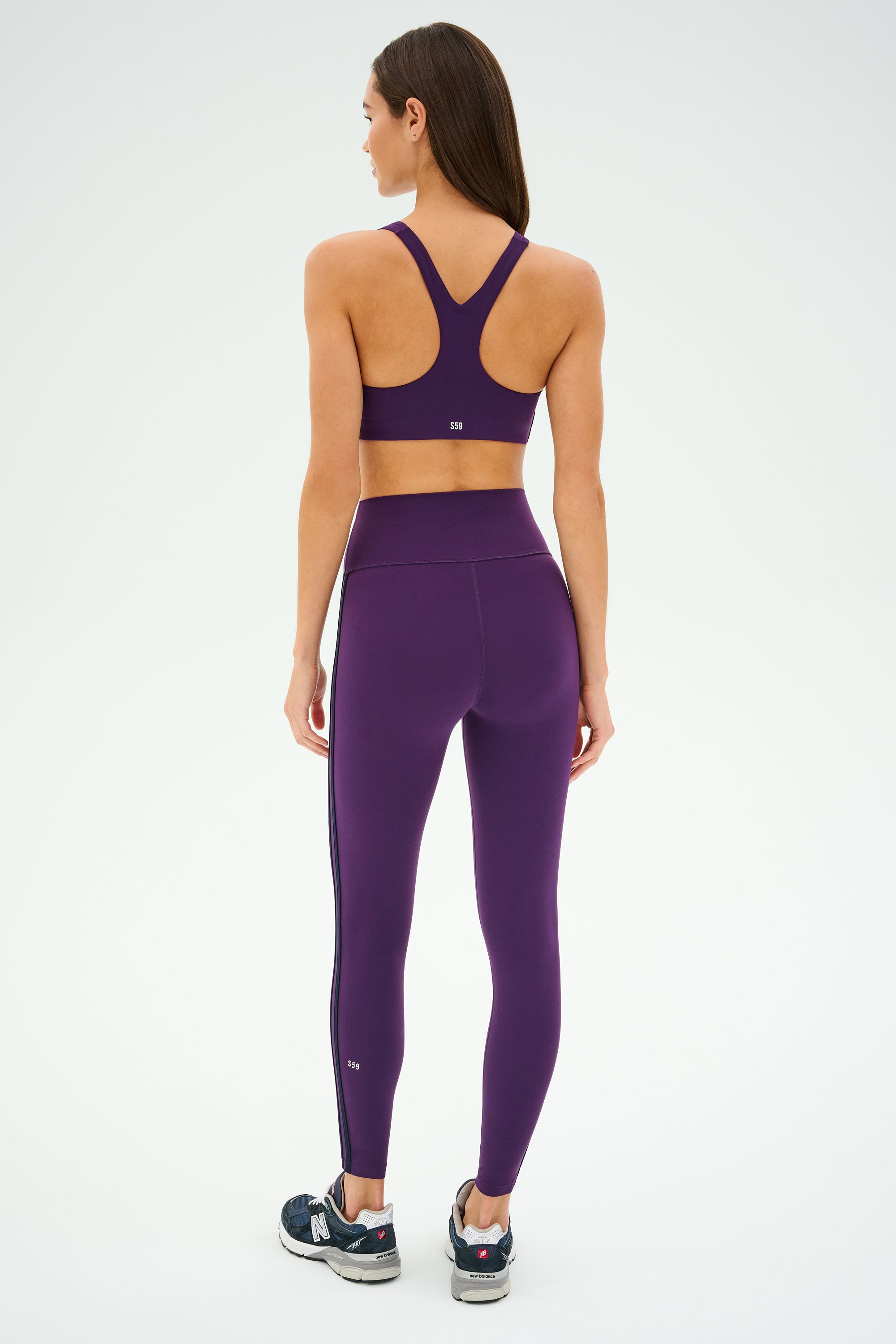 A person is wearing the Ella Airweight Bra in Black Currant/Indigo from SPLITS59 with matching leggings, standing with their back facing the camera, ready for a session of hot yoga.