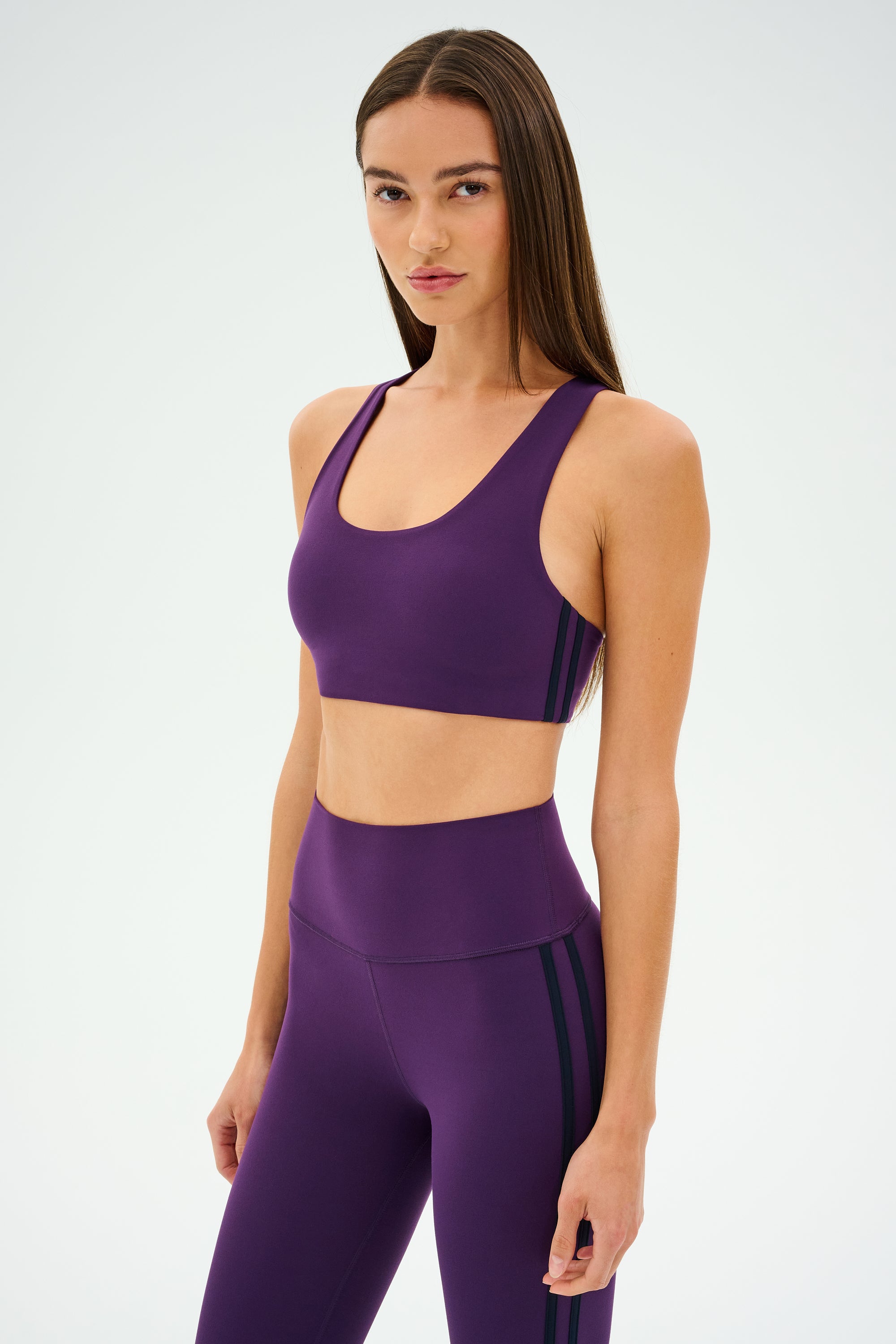 Wearing the Ella Airweight Bra in Black Currant/Indigo by SPLITS59, paired with matching leggings, a person confidently poses against a plain background, ready for hot yoga.