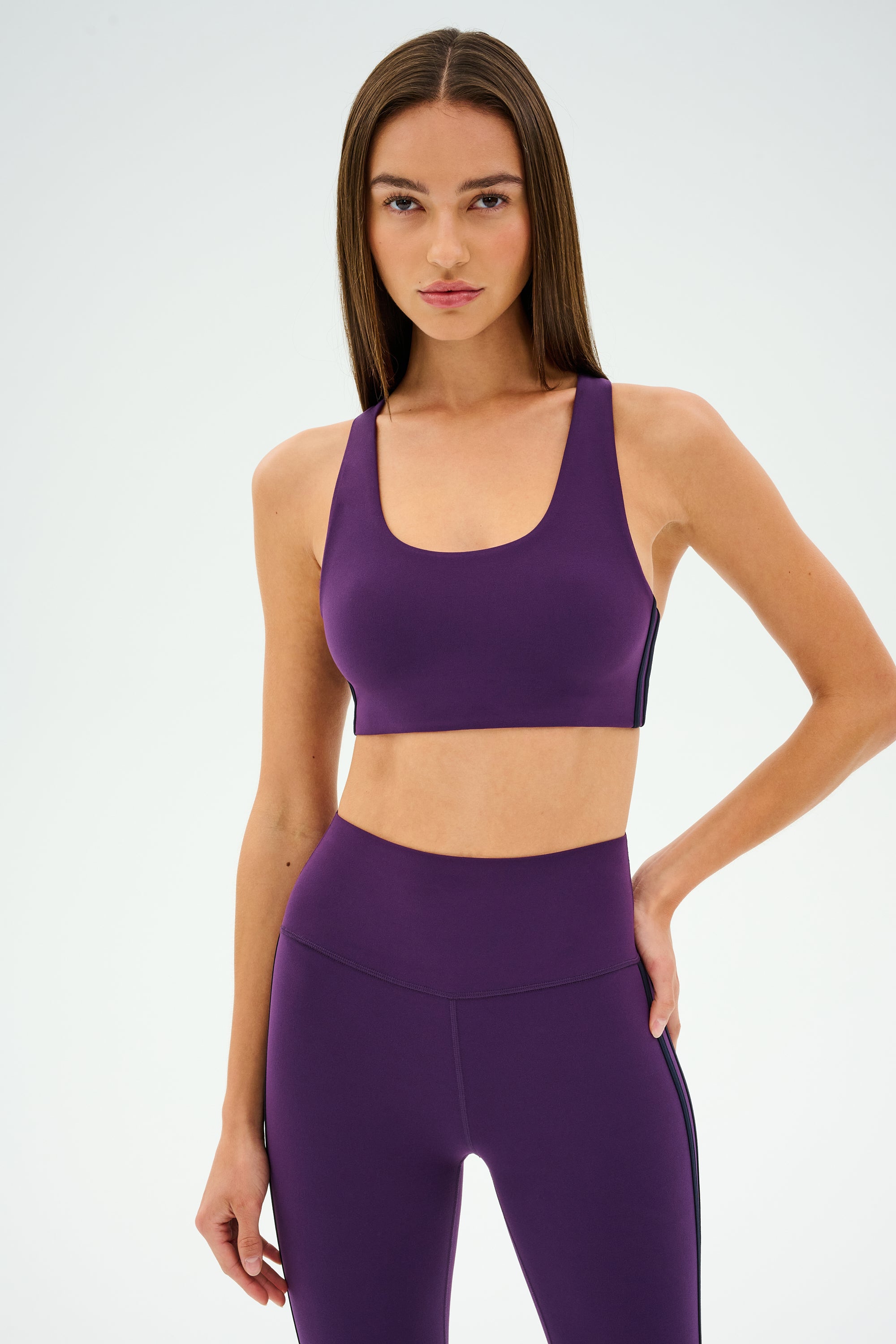 Wearing the SPLITS59 Ella Airweight Bra in Black Currant/Indigo, a person stands confidently against a plain background, ready for hot yoga.