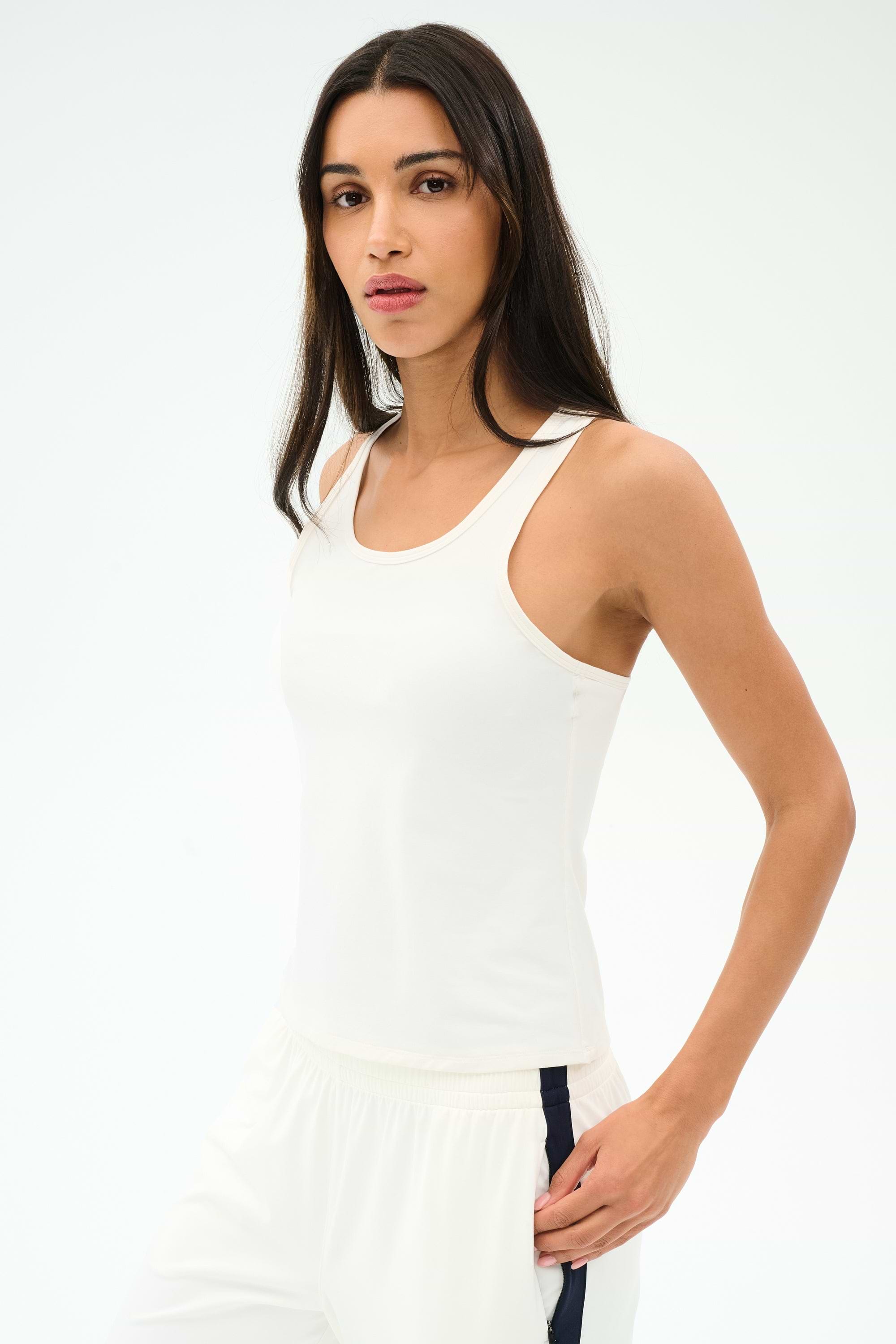 A woman with long dark hair wears the SPLITS59 Mel Airweight Lite Tank in white, paired with white pants featuring a navy stripe, standing against a plain light background.