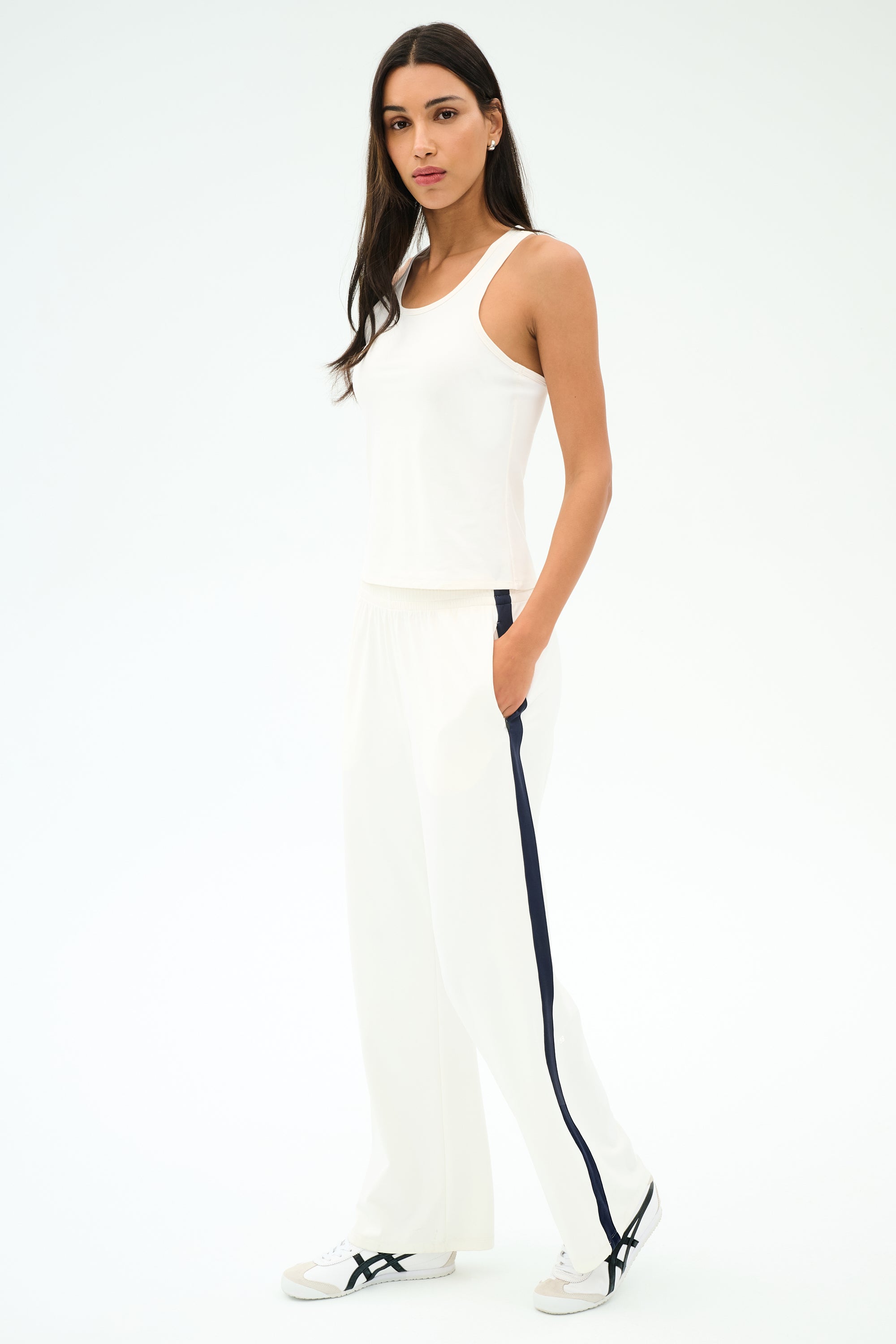 A woman poses with hands in pockets, dressed in the SPLITS59 Mel Airweight Lite Tank in white, wide-leg white pants featuring a black side stripe, and white sneakers against a plain background.
