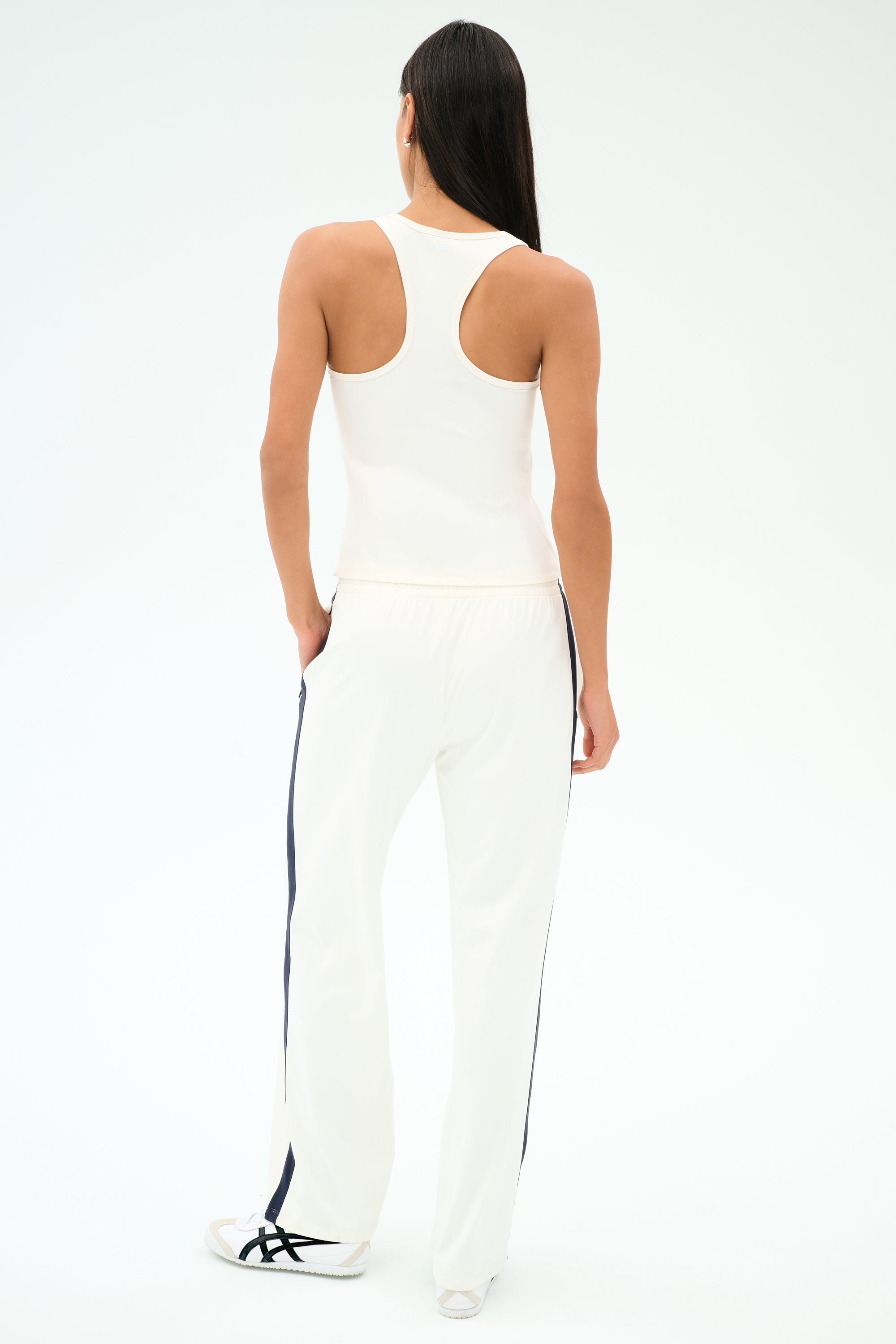 A person stands facing away against a plain white background, wearing the SPLITS59 Mel Airweight Lite Tank in white and white pants with black side stripes.