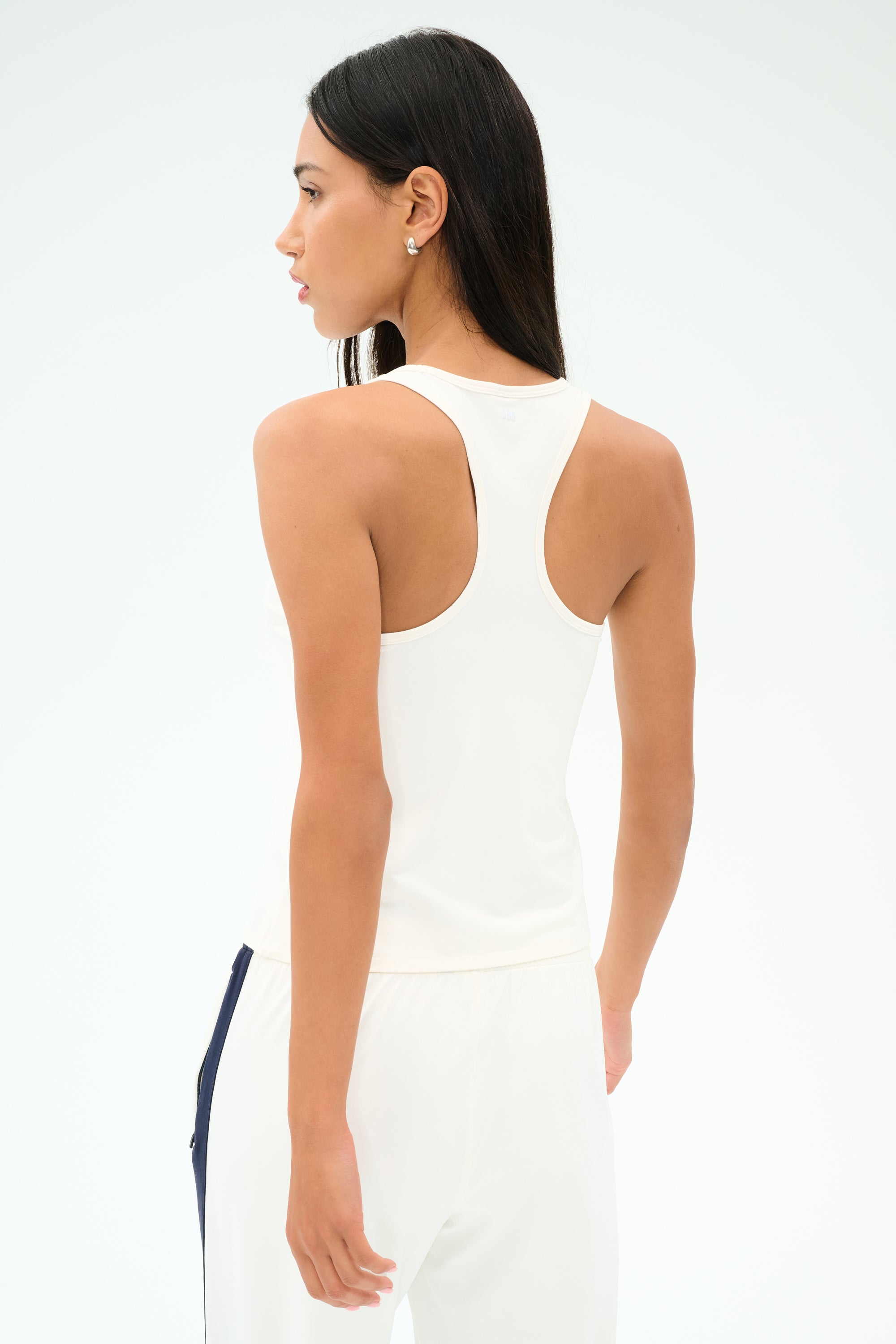 A person with long dark hair wears the SPLITS59 Mel Airweight Lite Tank in white, paired with white pants, standing facing away against a plain background.