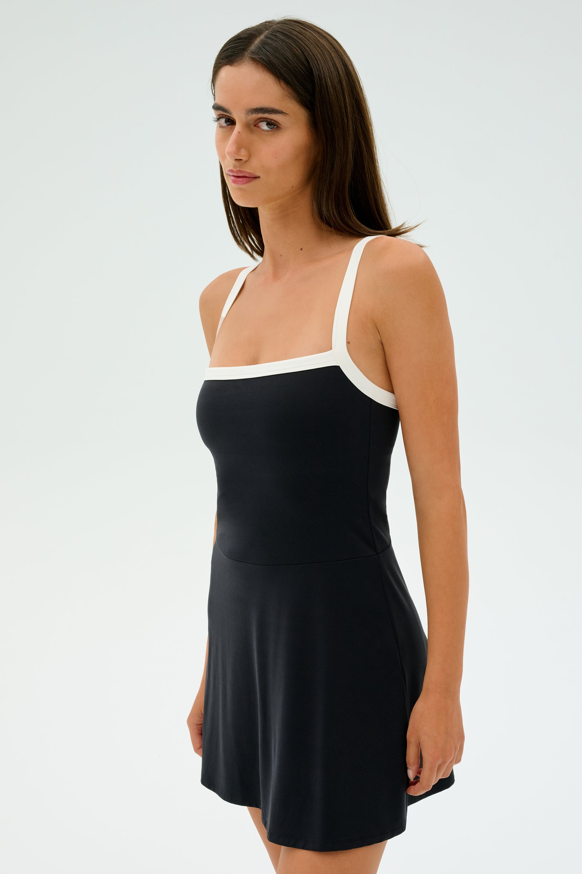Wearing the SPLITS59 Monah Rigor Dress in black with white straps, a woman stands against a plain background. The dress is crafted from a sleek blend of spandex and nylon as she looks slightly to the side.