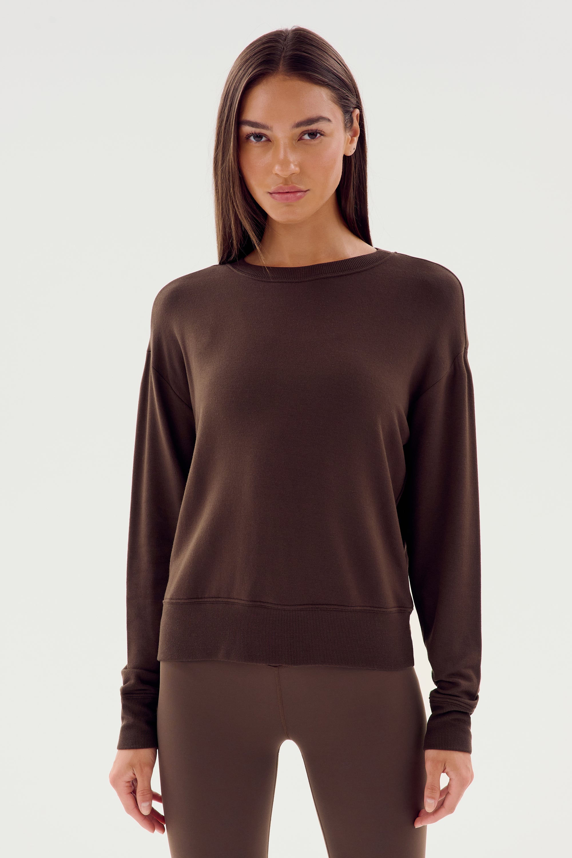 A woman with long straight hair stands facing the camera, wearing a plain dark long-sleeve top and matching leggings against a neutral background, showcasing the Sonja Fleece Sweatshirt in Dark Chocolate by SPLITS59.