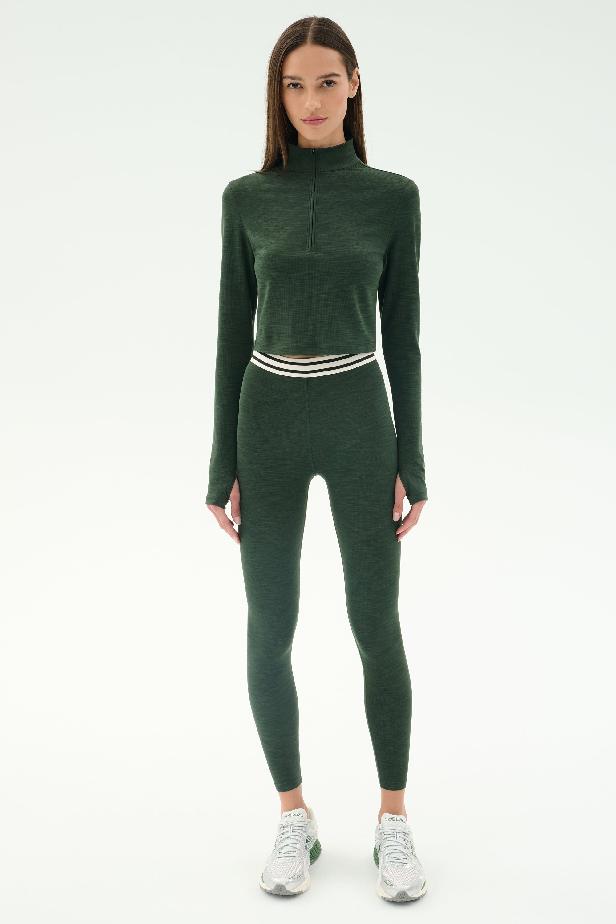 A woman faces forward wearing SPLITS59’s Bailey Active Rib High Waist 7/8 leggings in Heather Military with a matching dark green zip-up athletic top and white sneakers, set against a white background.