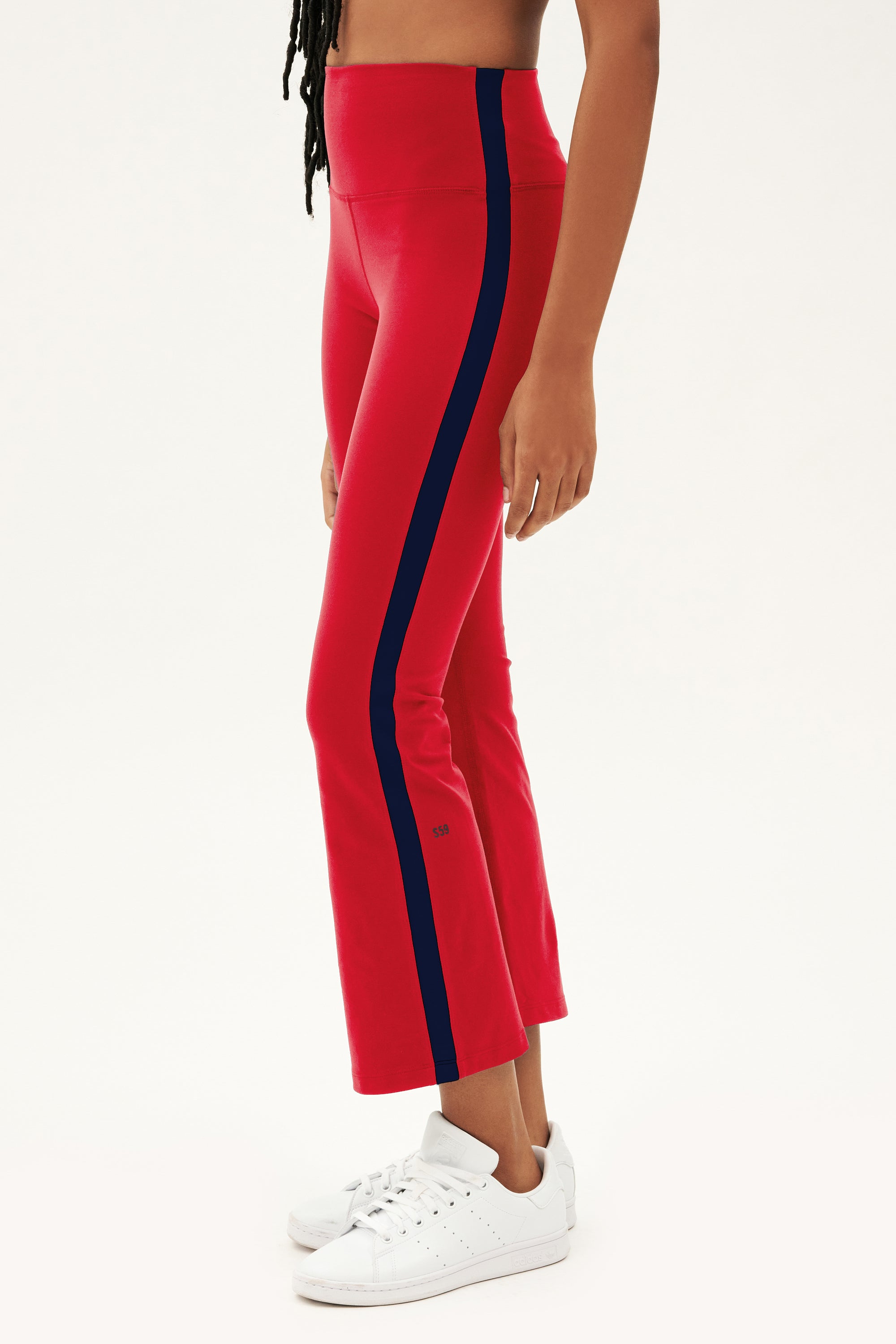 A person wears SPLITS59 Raquel High Waist Crop leggings in Pirate Red/Indigo, styled with white sneakers, standing before a plain white background.