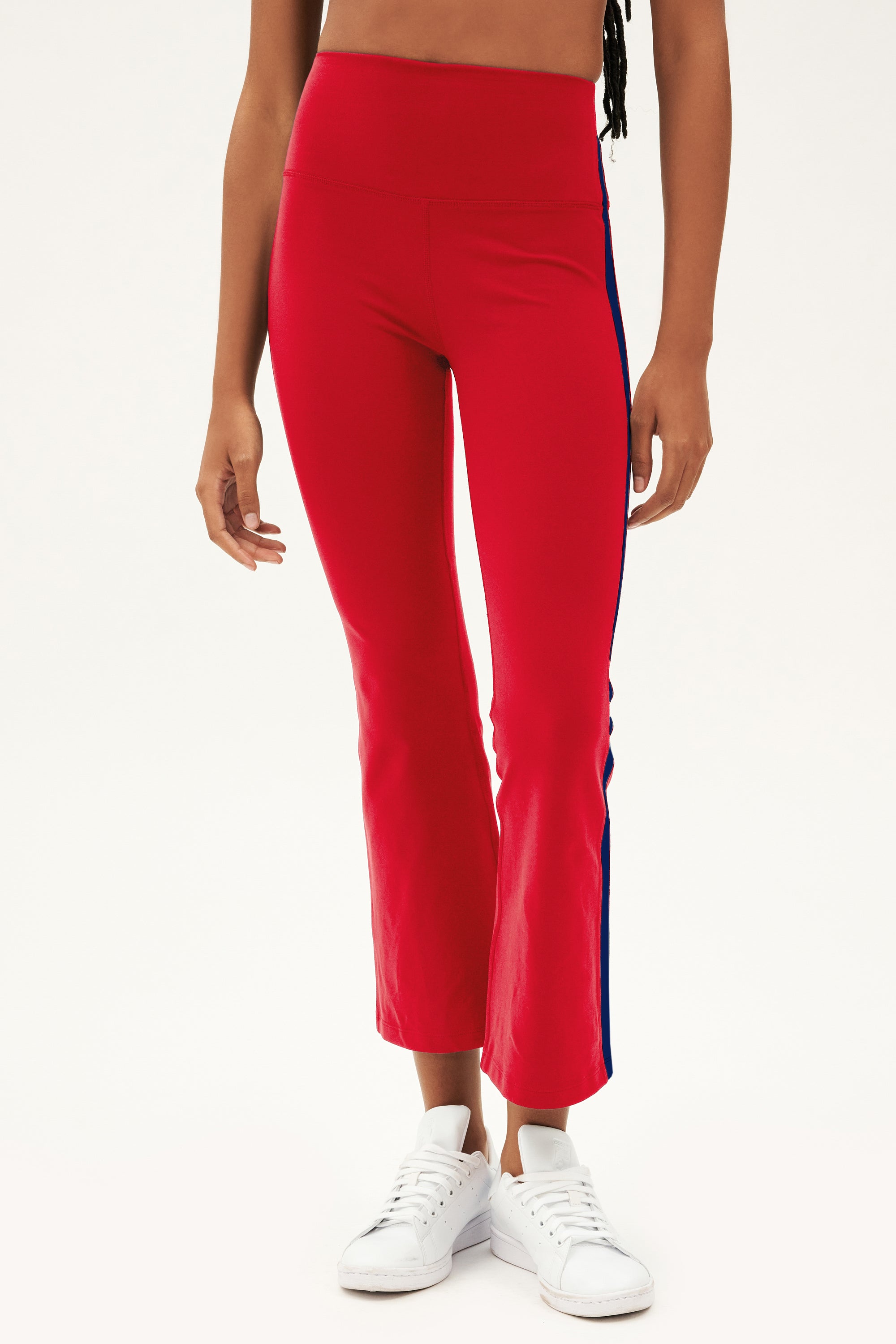 A person sports the SPLITS59 Raquel High Waist Crop in Pirate Red/Indigo, featuring a blue side stripe and 4-way stretch fabric, paired with white sneakers—ideal for yoga or everyday wear.