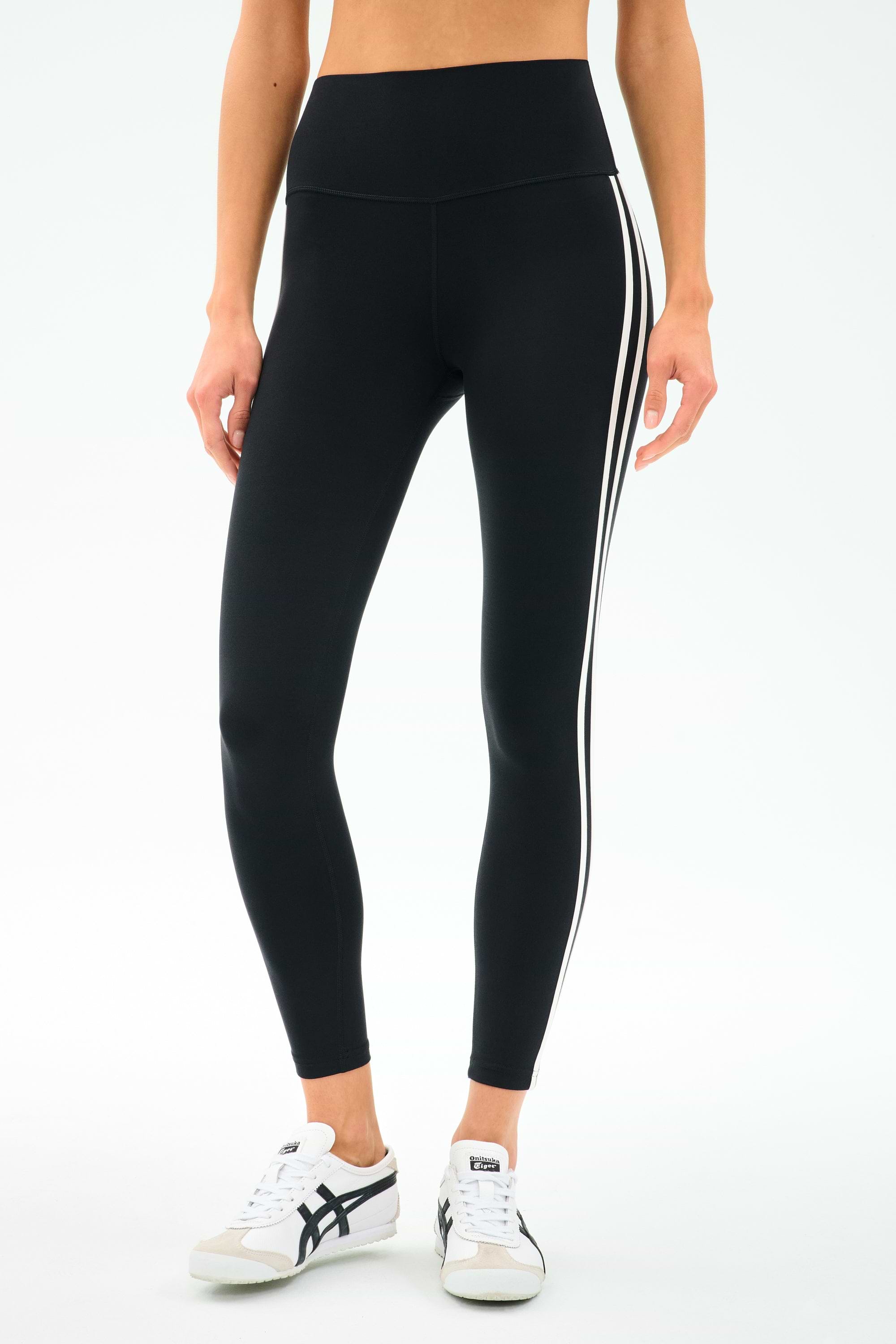 Someone in SPLITS59 Ella High Waist Airweight 7/8 leggings in Black/White with side stripes and white sneakers stands against a light background—ideal for hot yoga or everyday wear.