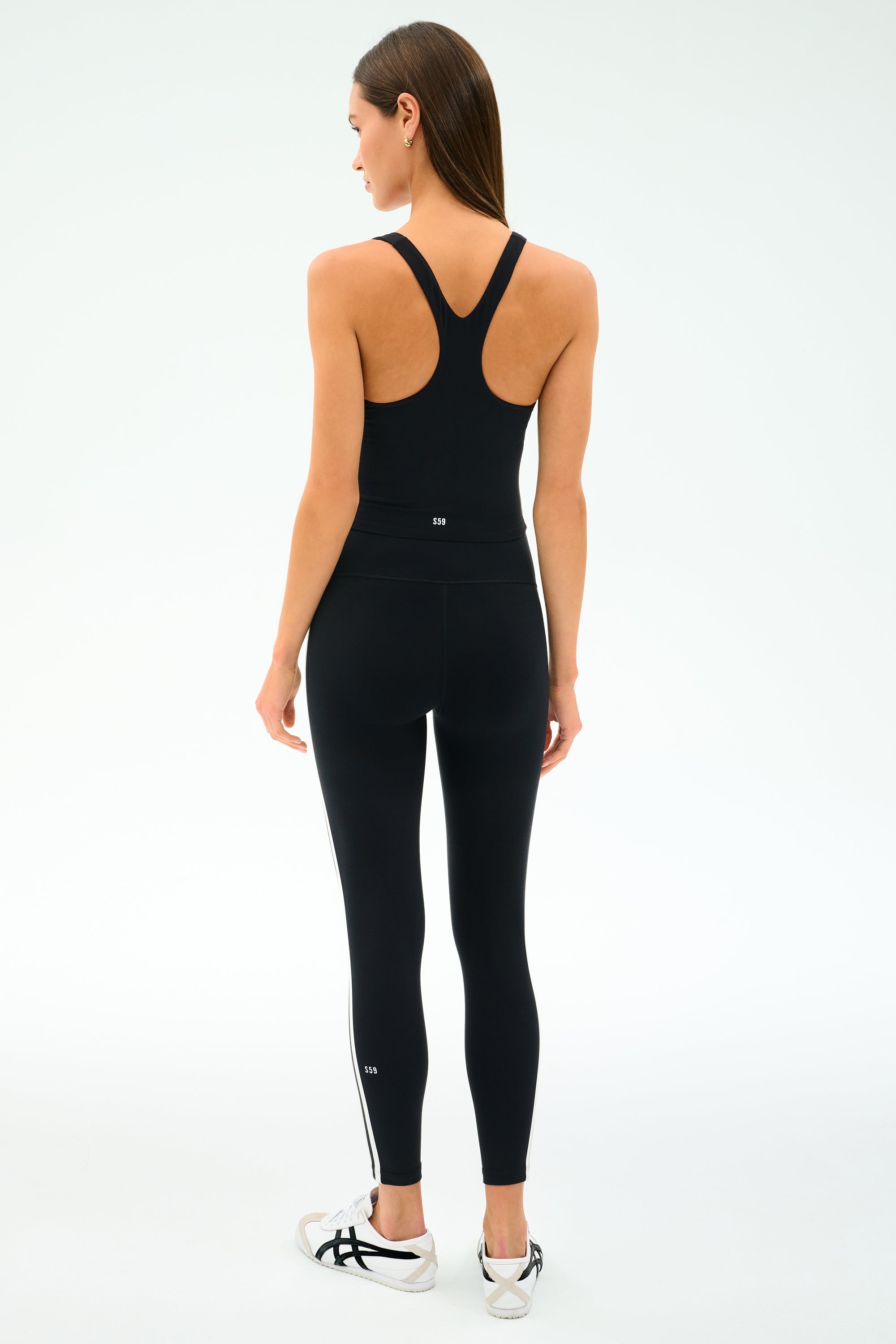 A woman stands facing away, wearing the SPLITS59 Ella High Waist Airweight 7/8 leggings in Black/White and a black racerback tank top, paired with white sneakers—ideal attire for hot yoga sessions against a plain light background.
