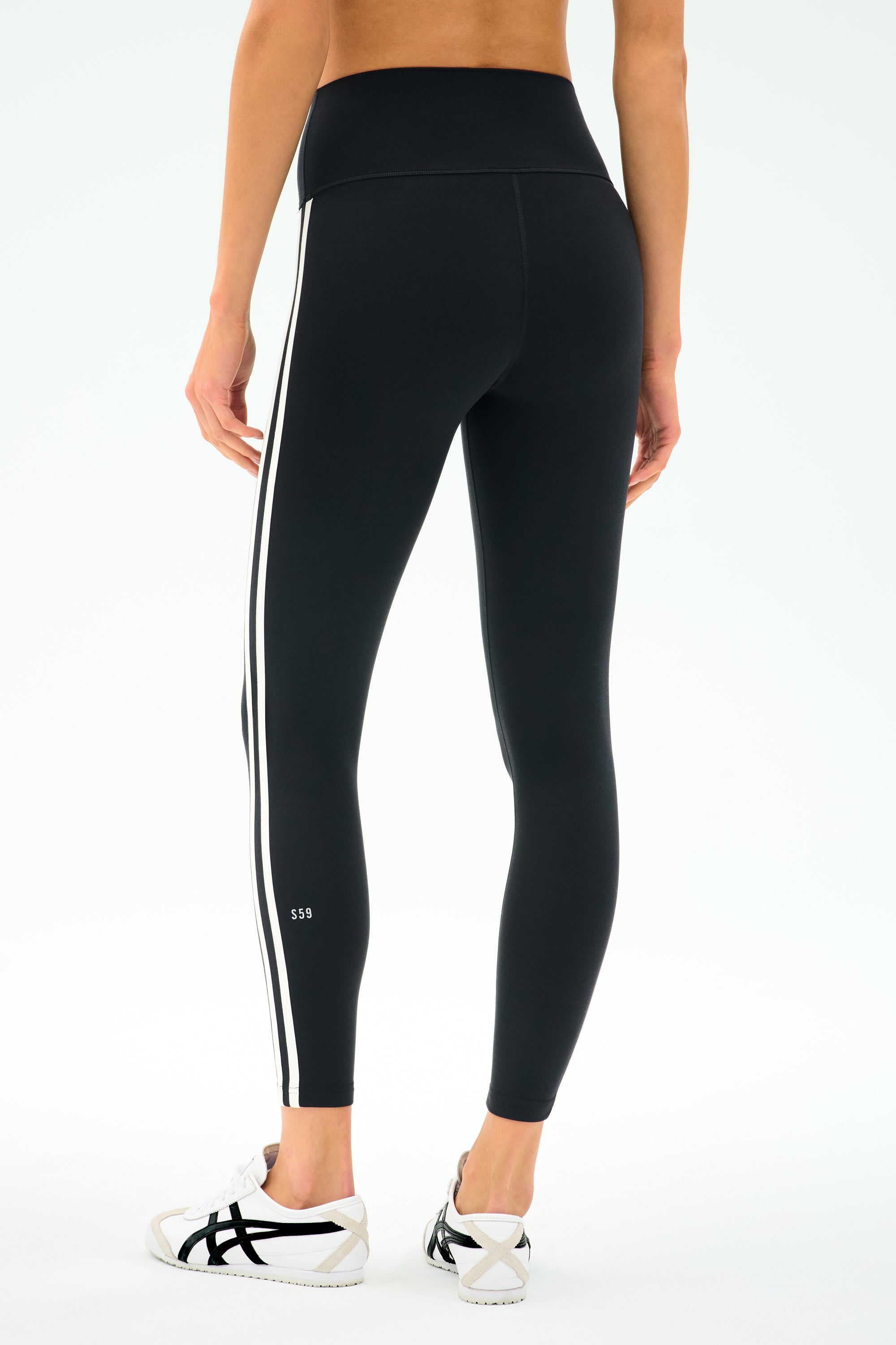 Person wears SPLITS59 Ella High Waist Airweight 7/8 Legging in Black/White—featuring white side stripes—paired with white athletic shoes, perfect for hot yoga, standing against a plain light background.