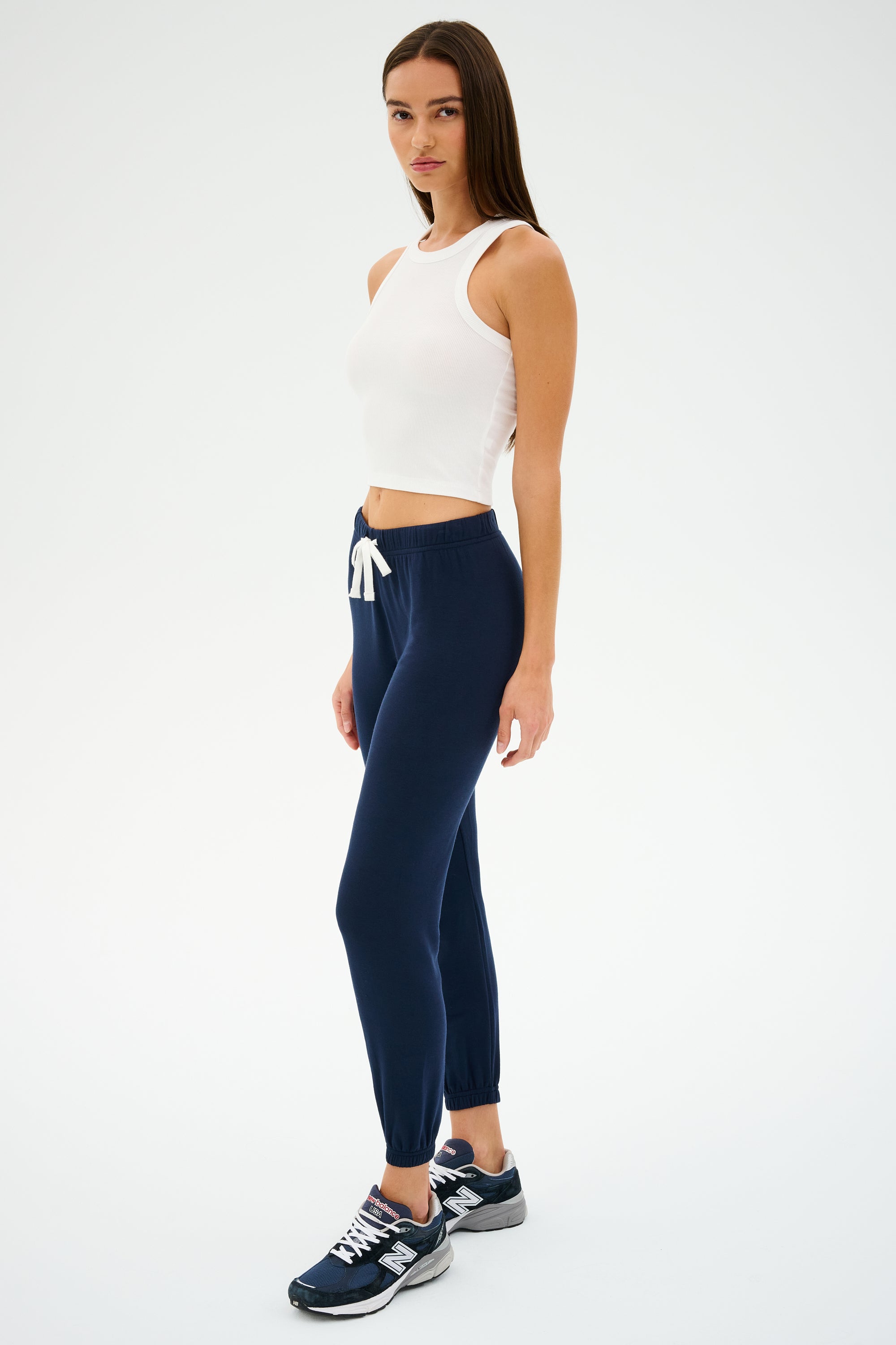 A person wearing the SPLITS59 Kiki Rib Crop Tank in white, expertly designed from baby rib fabric, pairs it with navy joggers and sneakers while standing on a plain background.