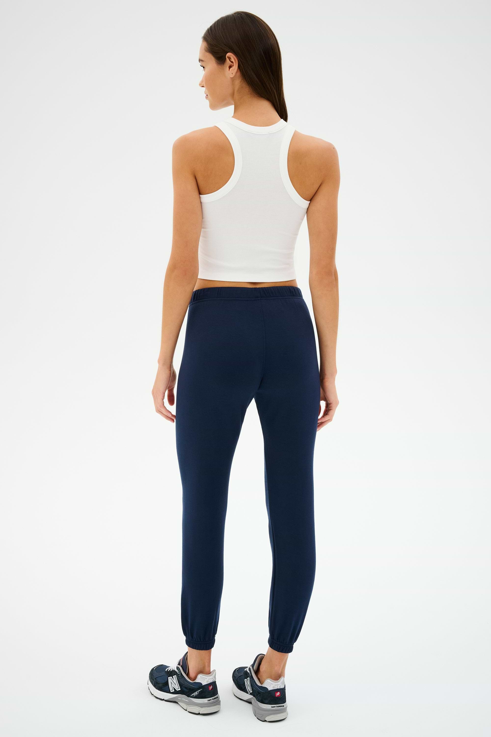 A person stands with their back to the camera, wearing a Kiki Rib Crop Tank in white from SPLITS59, navy joggers, and black sneakers against a plain white background.