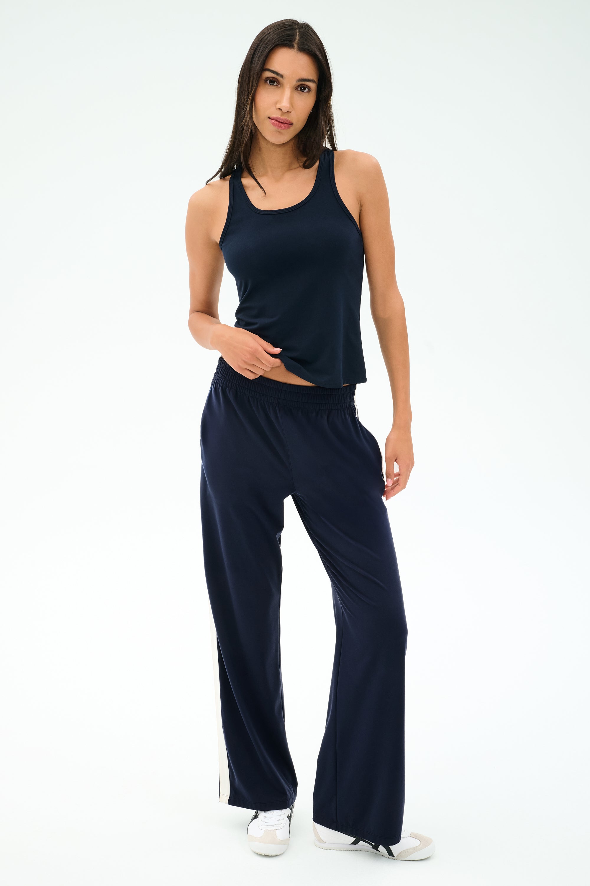 A woman stands against a plain background wearing the SPLITS59 Mel Airweight Lite Tank in Indigo, matching wide-leg pants with a white stripe, and white sneakers.