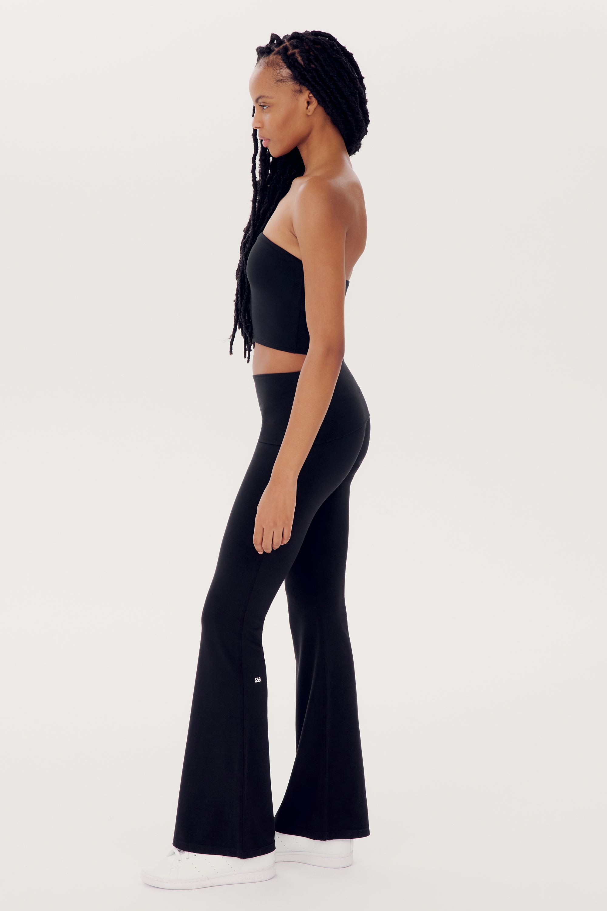 A woman with long braided hair stands in profile, wearing a black SPLITS59 Hazel Airweight Crop Bandeau - Black, black flared pants, and white sneakers.