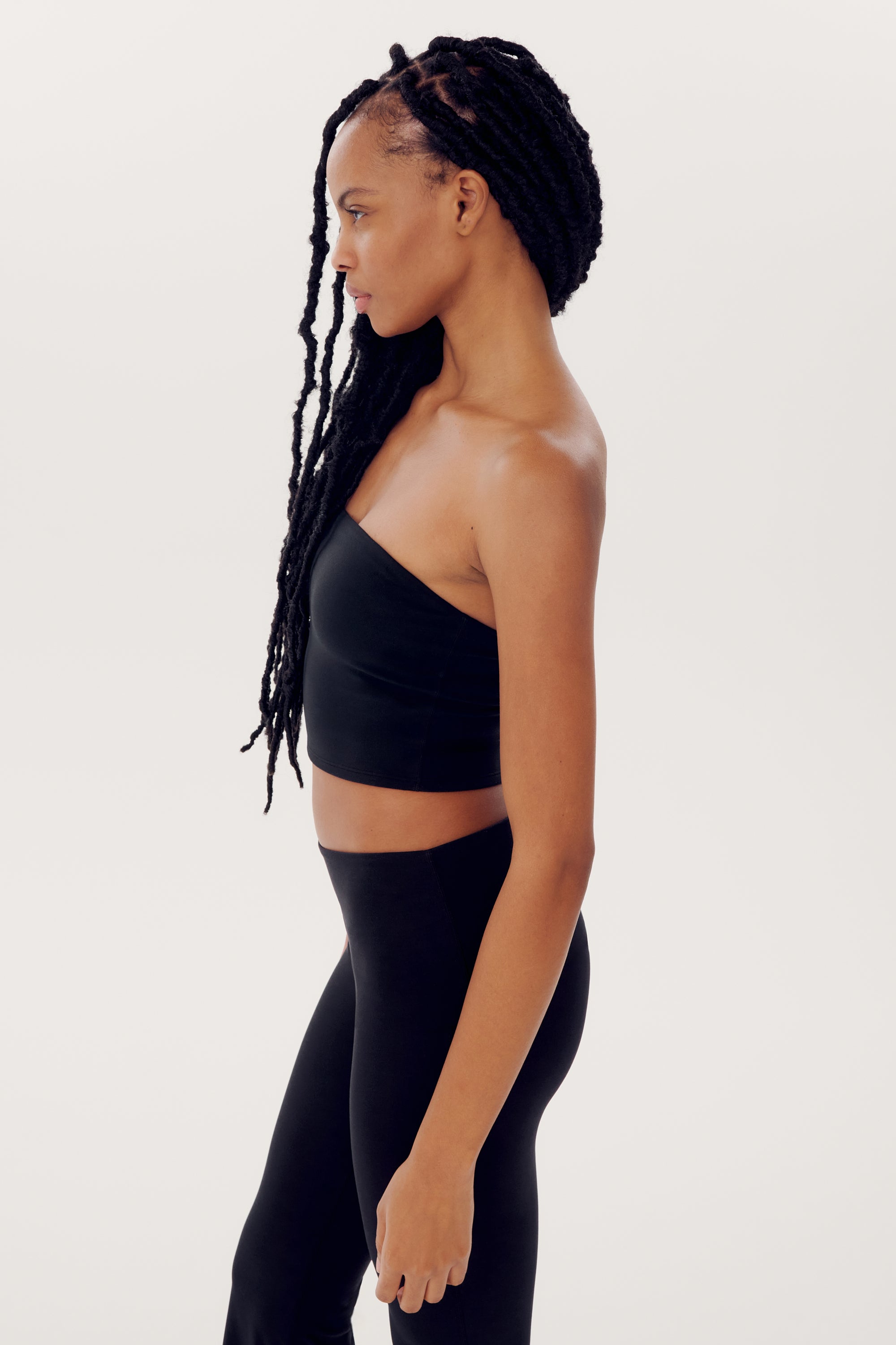 A person with long braided hair is standing in profile against a plain background, wearing a black SPLITS59 Hazel Airweight Crop Bandeau - Black and black nylon-spandex pants.