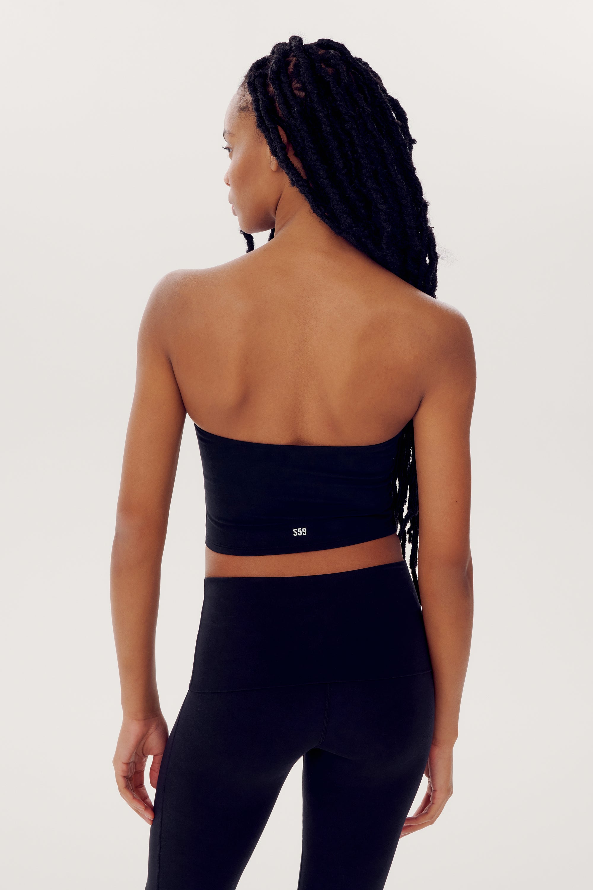 A person with long braided hair is shown from the back, wearing a Hazel Airweight Crop Bandeau - Black by SPLITS59 and black spandex leggings against a plain background.