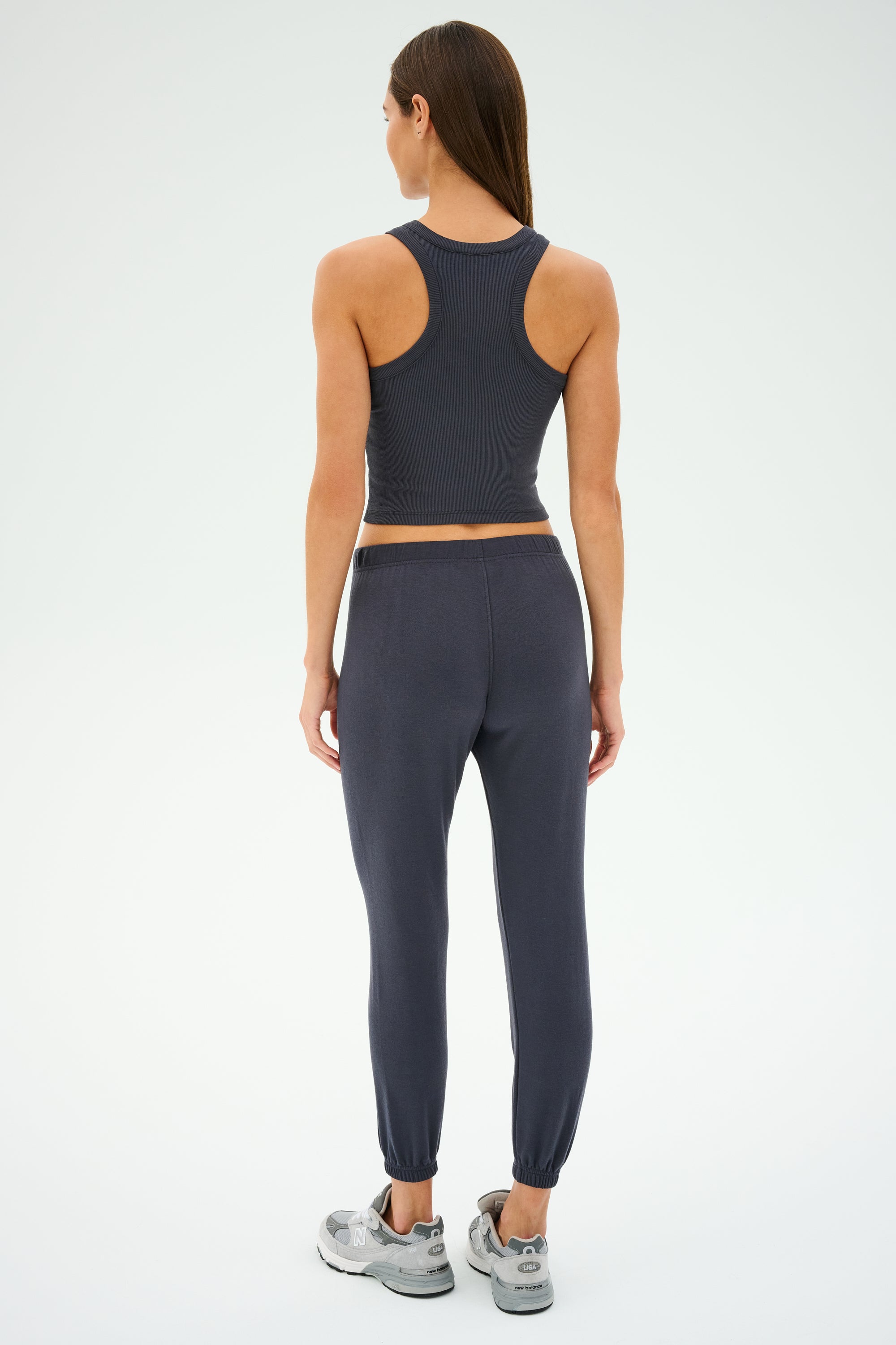 A woman stands facing away, showcasing the sleek SPLITS59 Kiki Rib Crop Tank in Graphite, paired with black joggers and gray sneakers against a plain backdrop. Her effortlessly chic look captures the MADE IN LOS ANGELES quality.