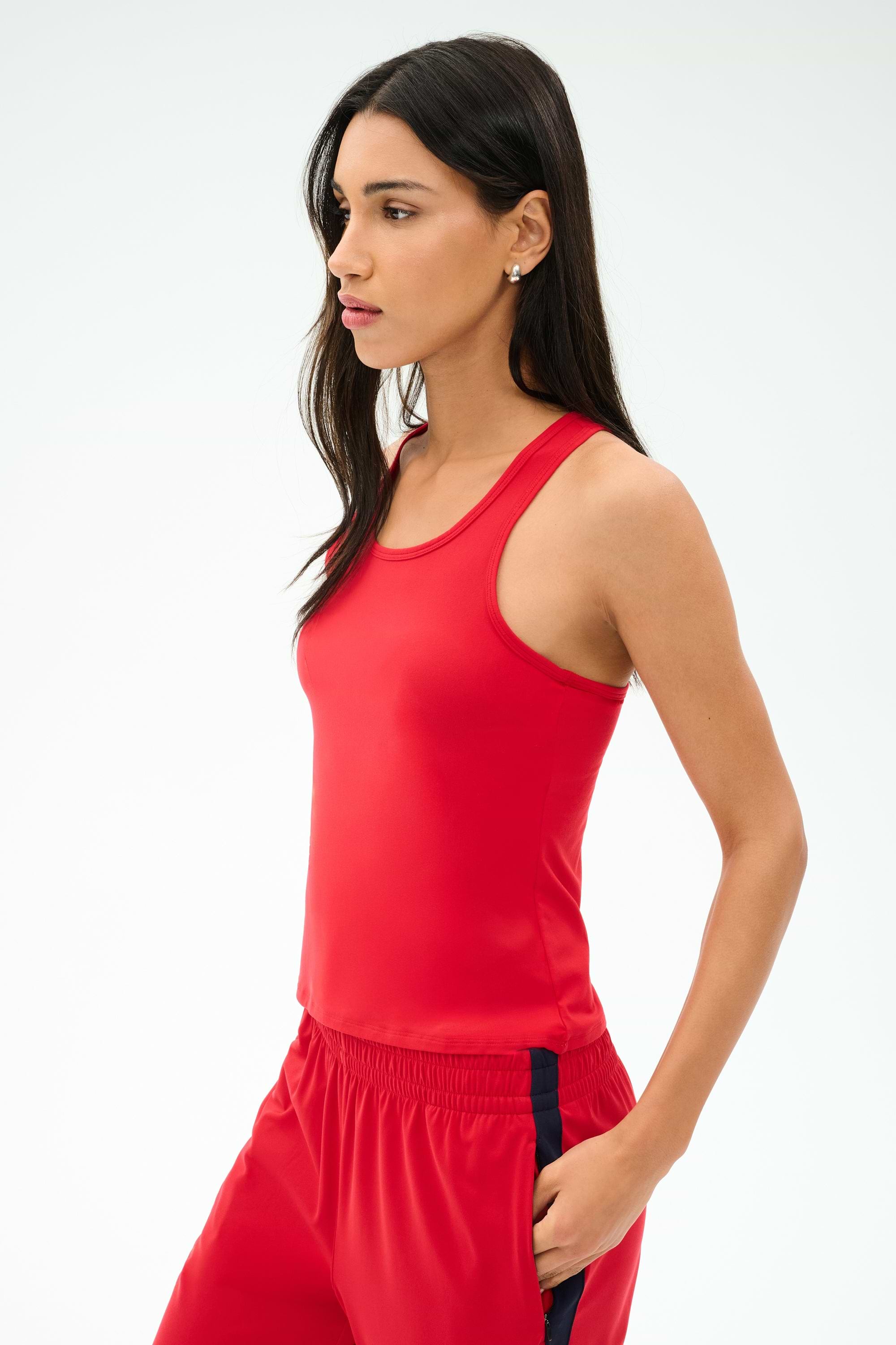 A woman wears the SPLITS59 Mel Airweight Lite Tank in Pirate Red with matching track pants featuring black stripes, standing facing left against a plain light background.