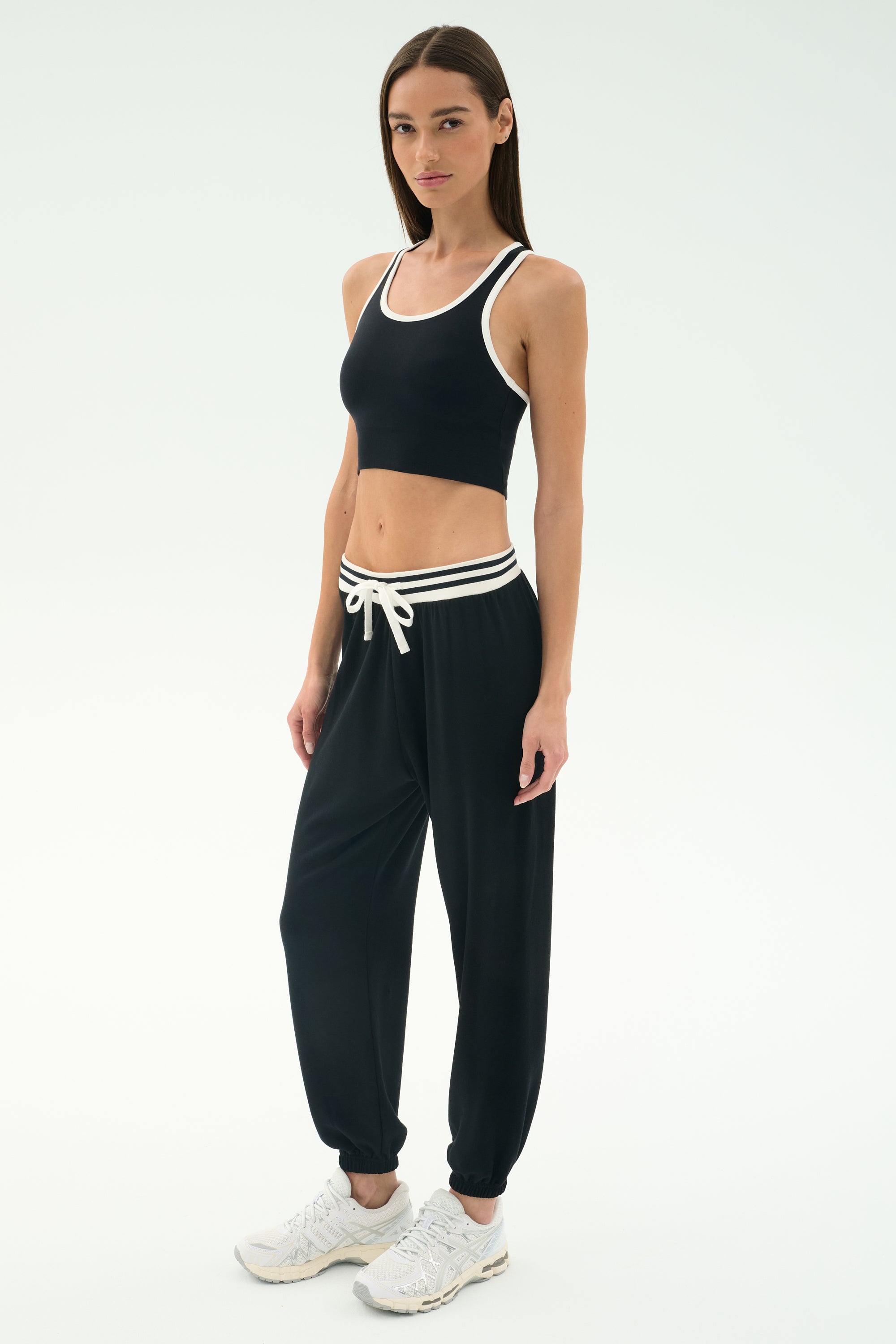 A woman stands in a studio wearing the SPLITS59 Andie Oversized Fleece Sweatpant W/ Stripe in black, paired with a black sports bra and white sneakers.