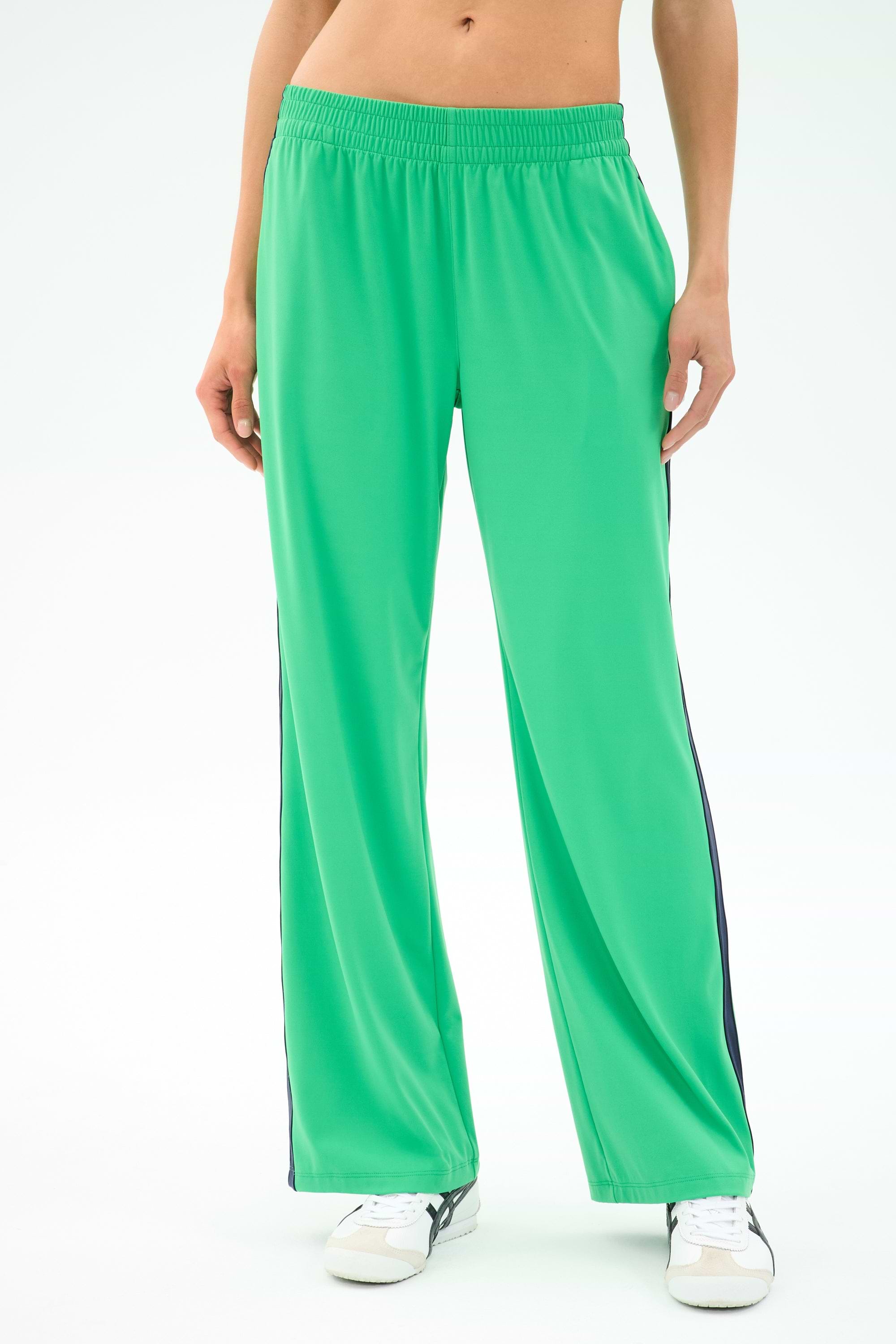 Person wearing SPLITS59 Maxie Rigor Trackpant in Grass/Indigo with black side stripes and white sneakers, standing against a plain background. Only the lower torso and legs are shown.