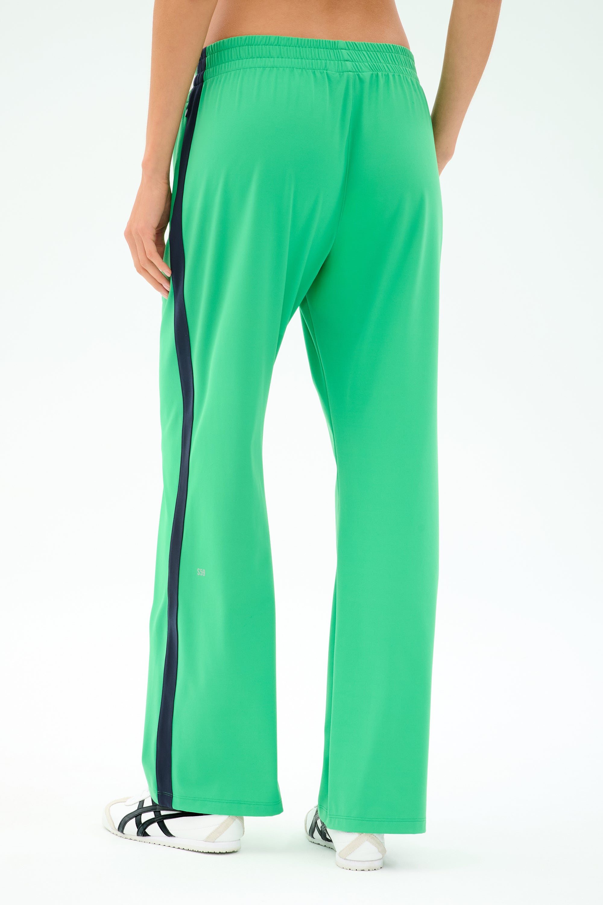 A person wears SPLITS59 Maxie Rigor Trackpant in Grass/Indigo, featuring a dark side stripe, paired with white sneakers and standing against a plain light background.