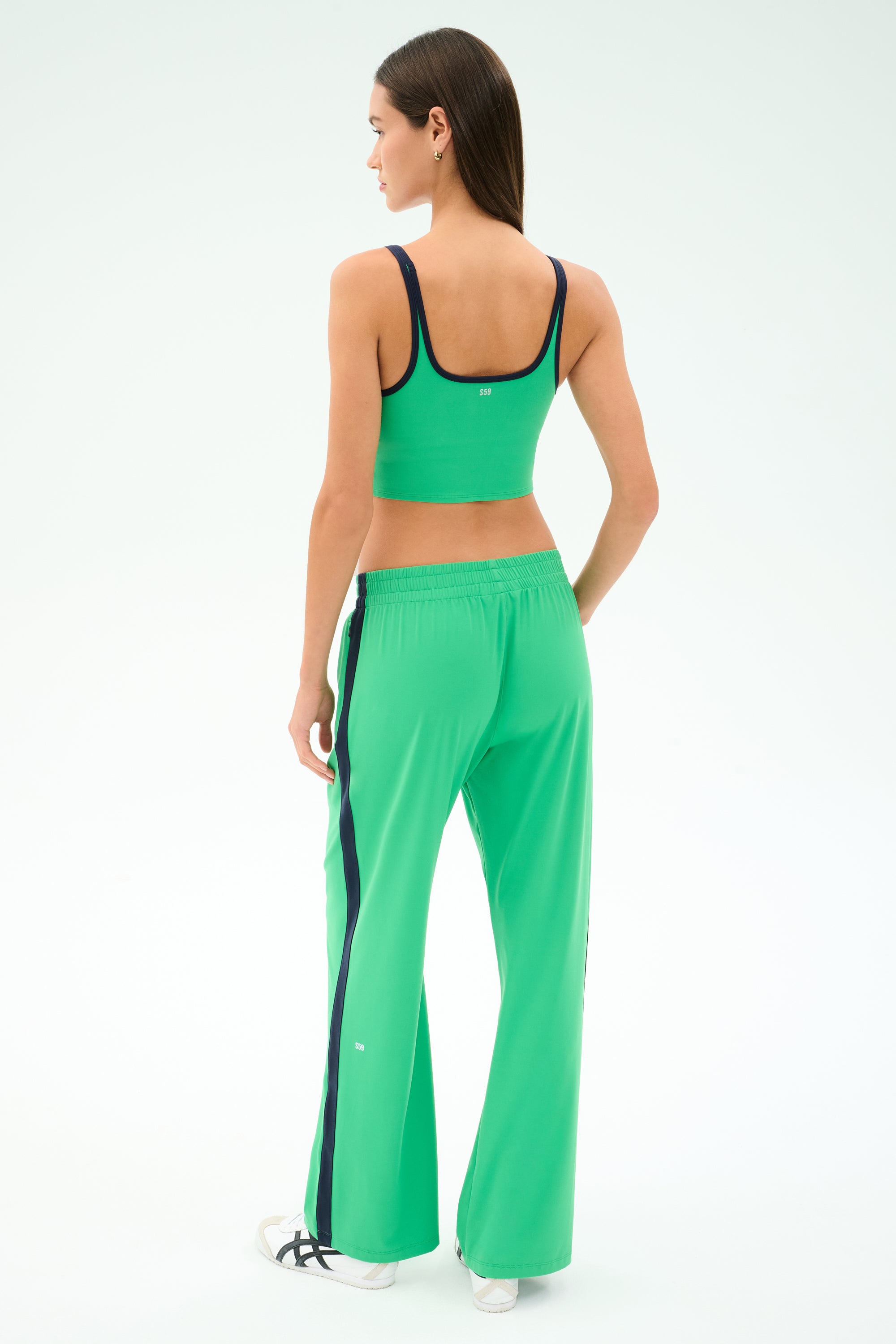 A woman faces away, wearing SPLITS59's Maxie Rigor Trackpant in Grass/Indigo with black stripes, paired with a green crop top and white sneakers.