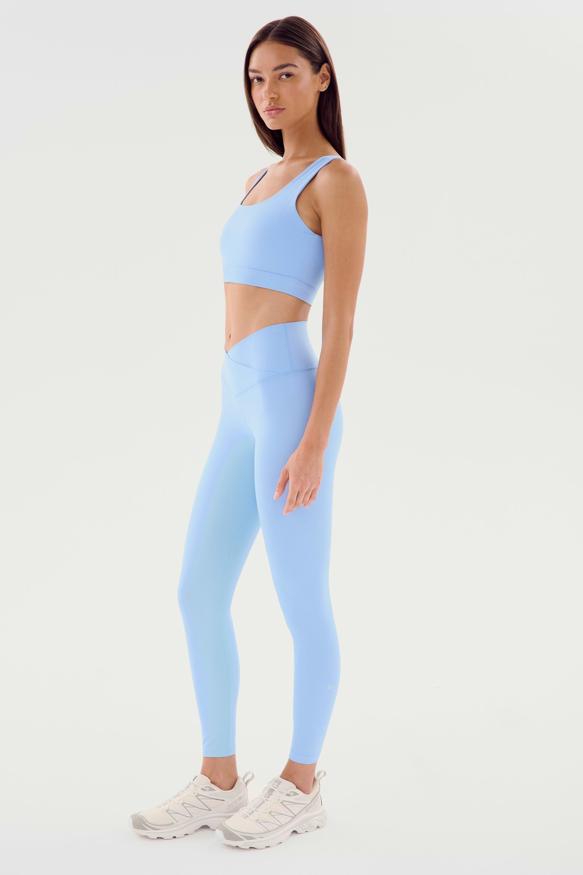 A woman stands against a white background wearing a light blue Sprint Rigor Bra - Skylight by SPLITS59 and leggings with white athletic shoes, ideal for gym workouts. She faces slightly to the right with her hands by her sides.