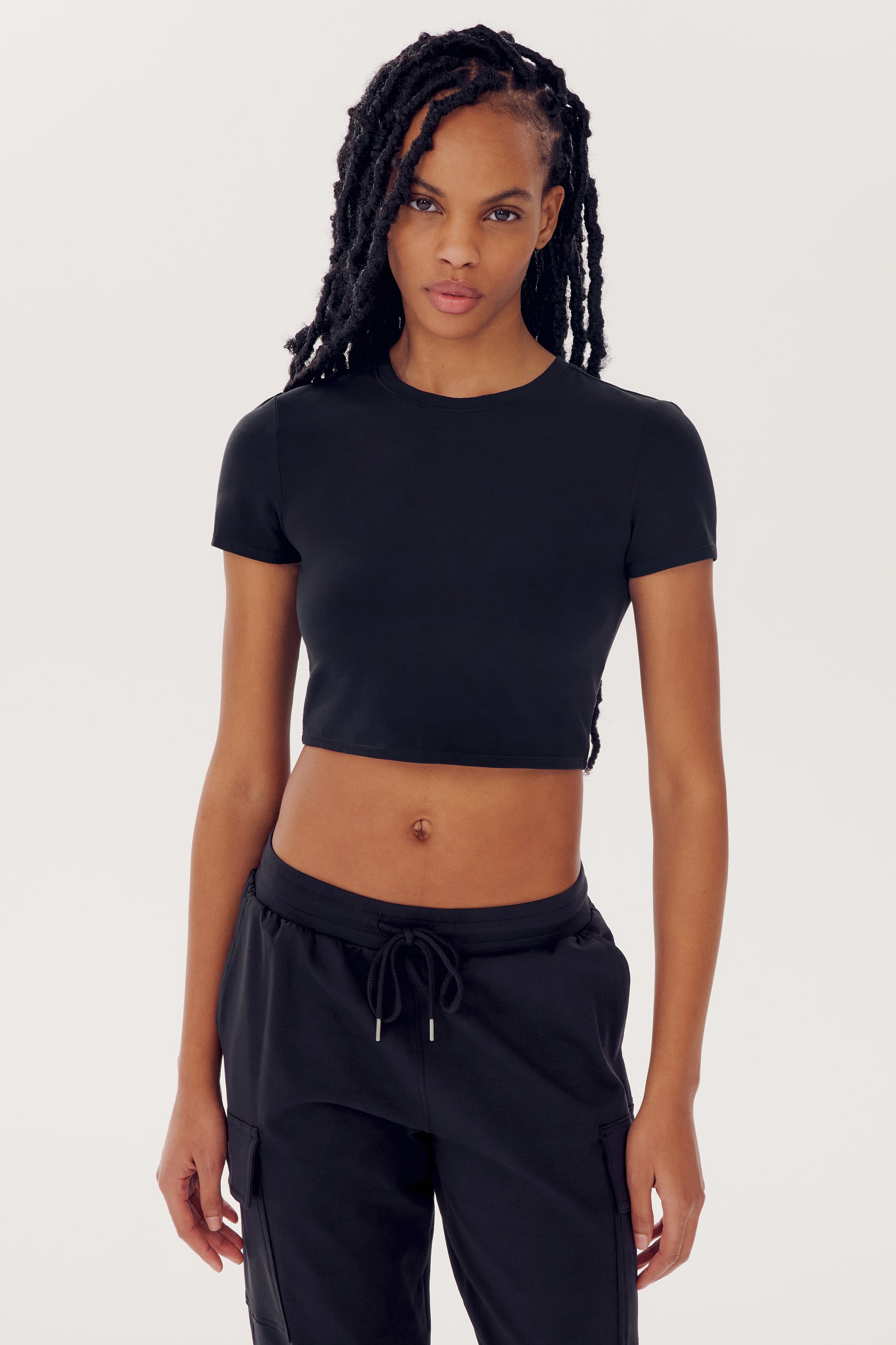 Person with long dreadlocks wearing a SPLITS59 Airweight S/S Crop - Black and black nylon pants, standing against a plain white background.