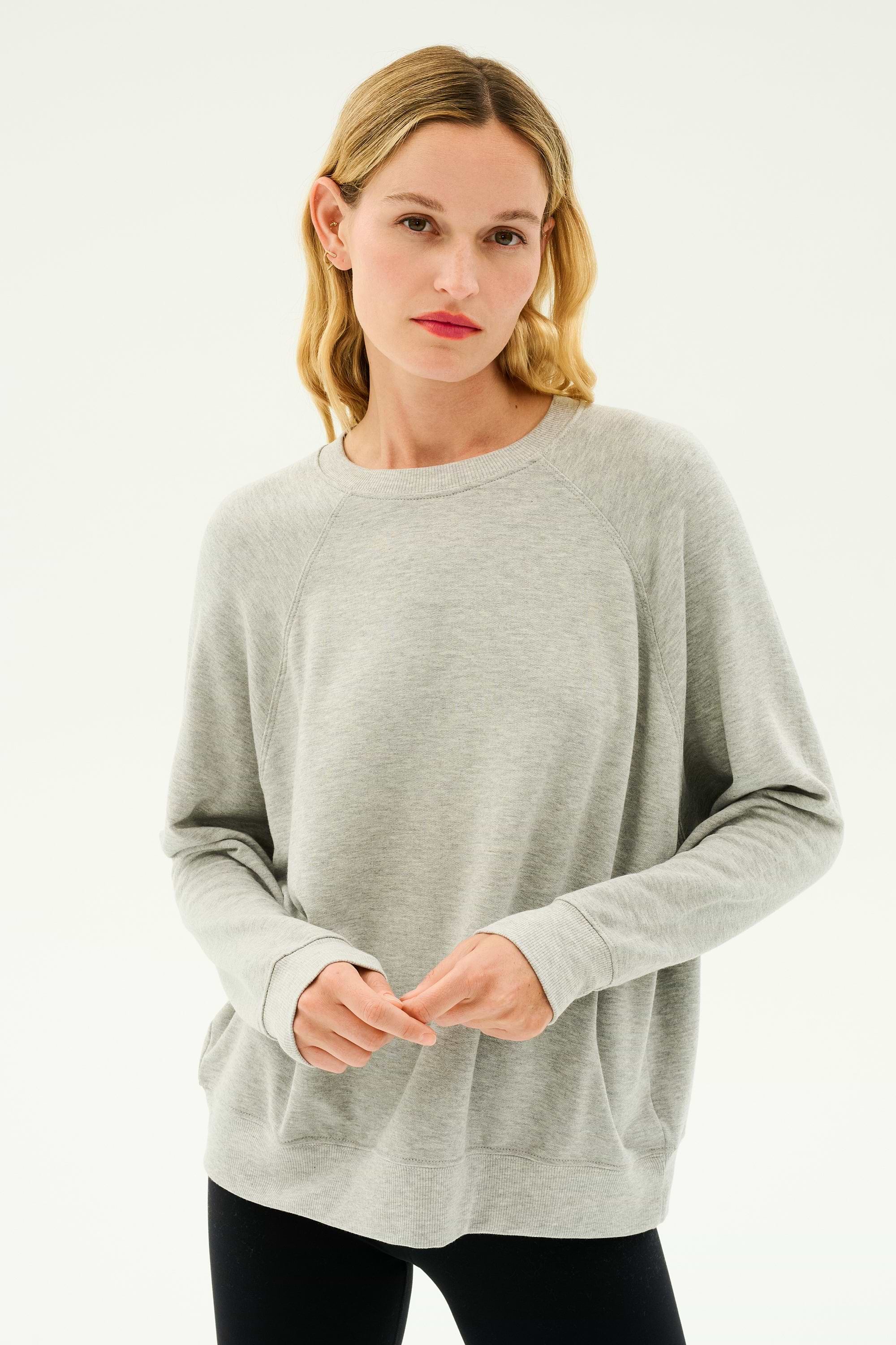 A person with blonde hair wearing a light gray Andie Fleece Sweatshirt - Heather Grey by SPLITS59 and black pants stands against a plain white background.