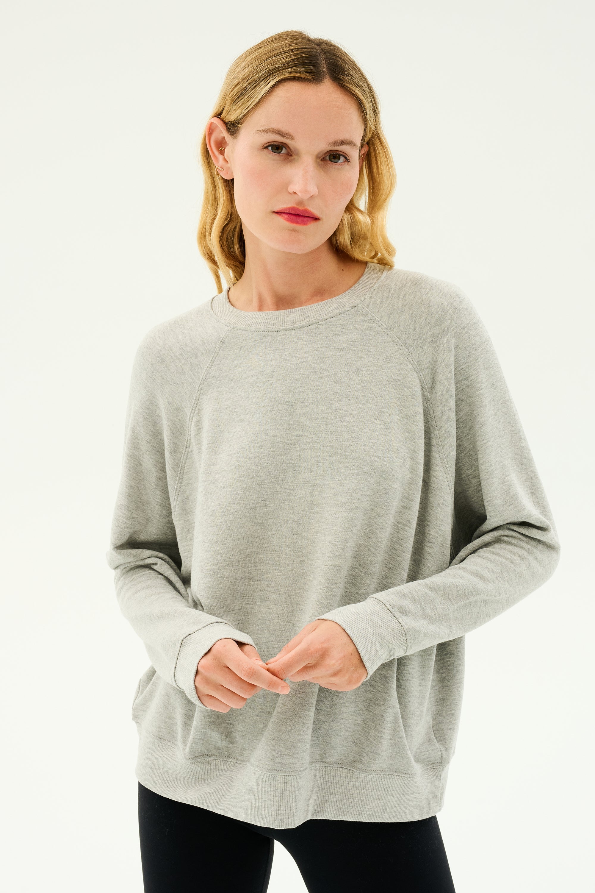 A person with blonde hair wearing a light gray Andie Fleece Sweatshirt - Heather Grey by SPLITS59 and black pants stands against a plain white background.